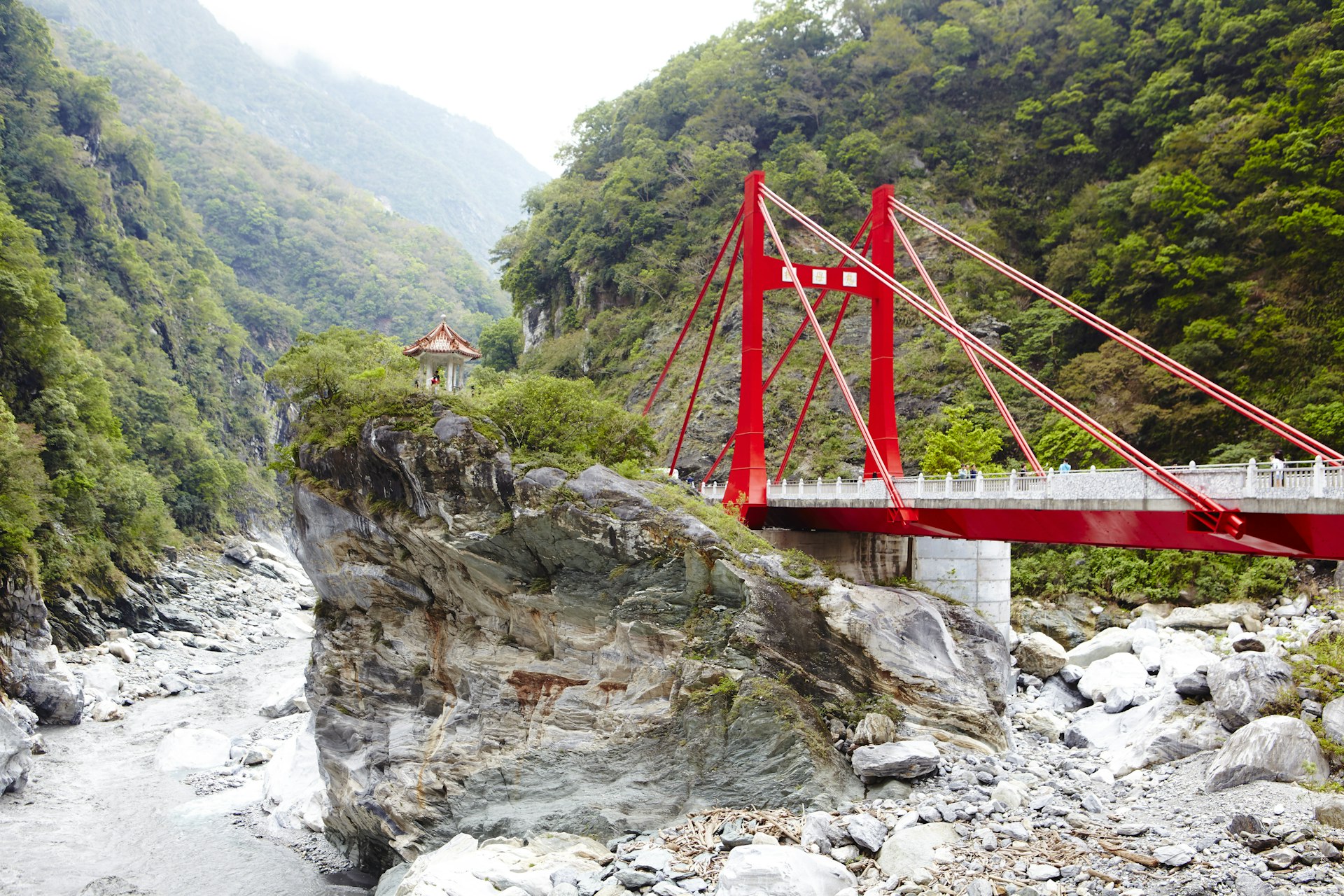 A red iron bridge leads to a small riverside pavilion