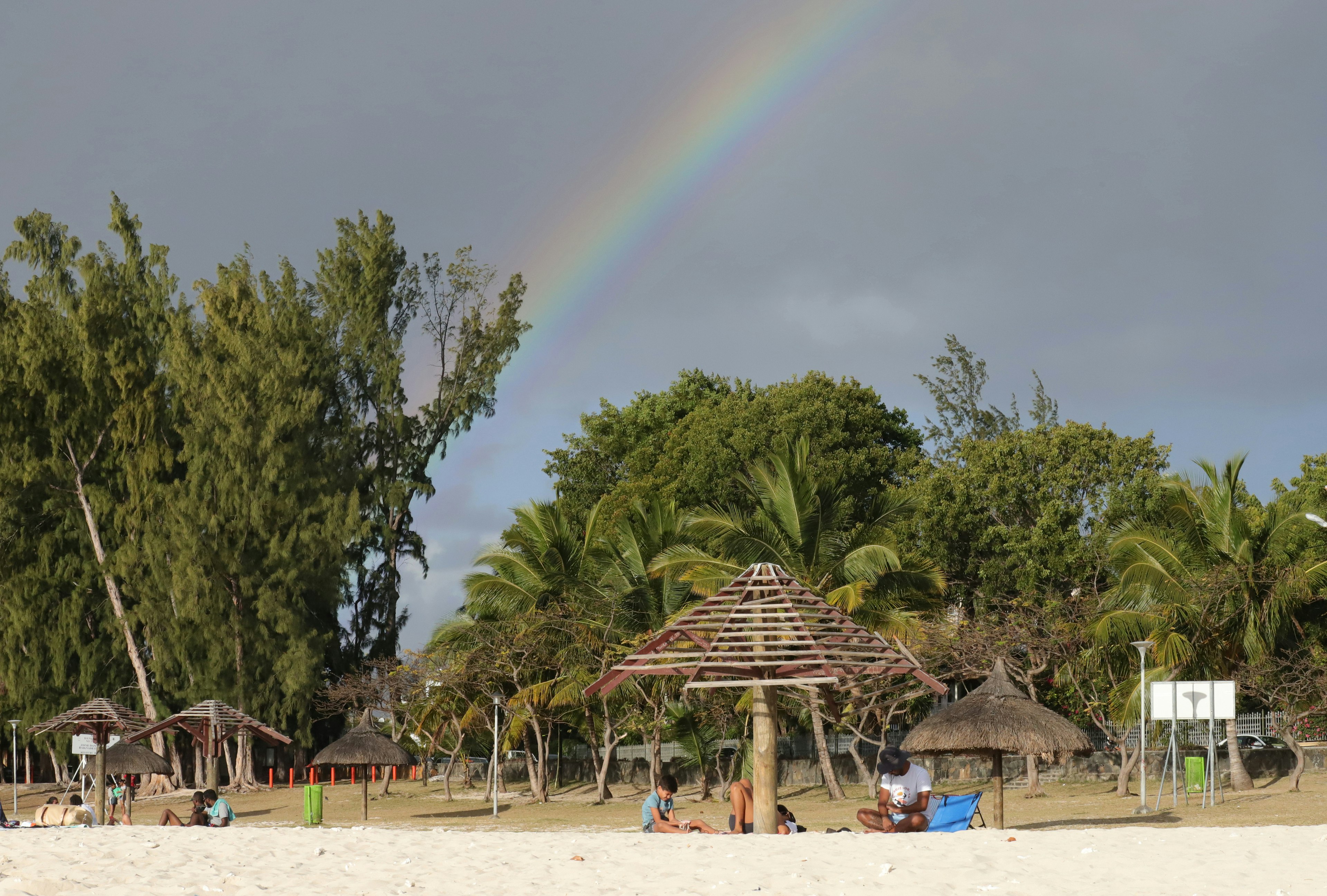 People relax in the shade of a wooden umbrella on the beach with a rainbow streaking across the sky behind them