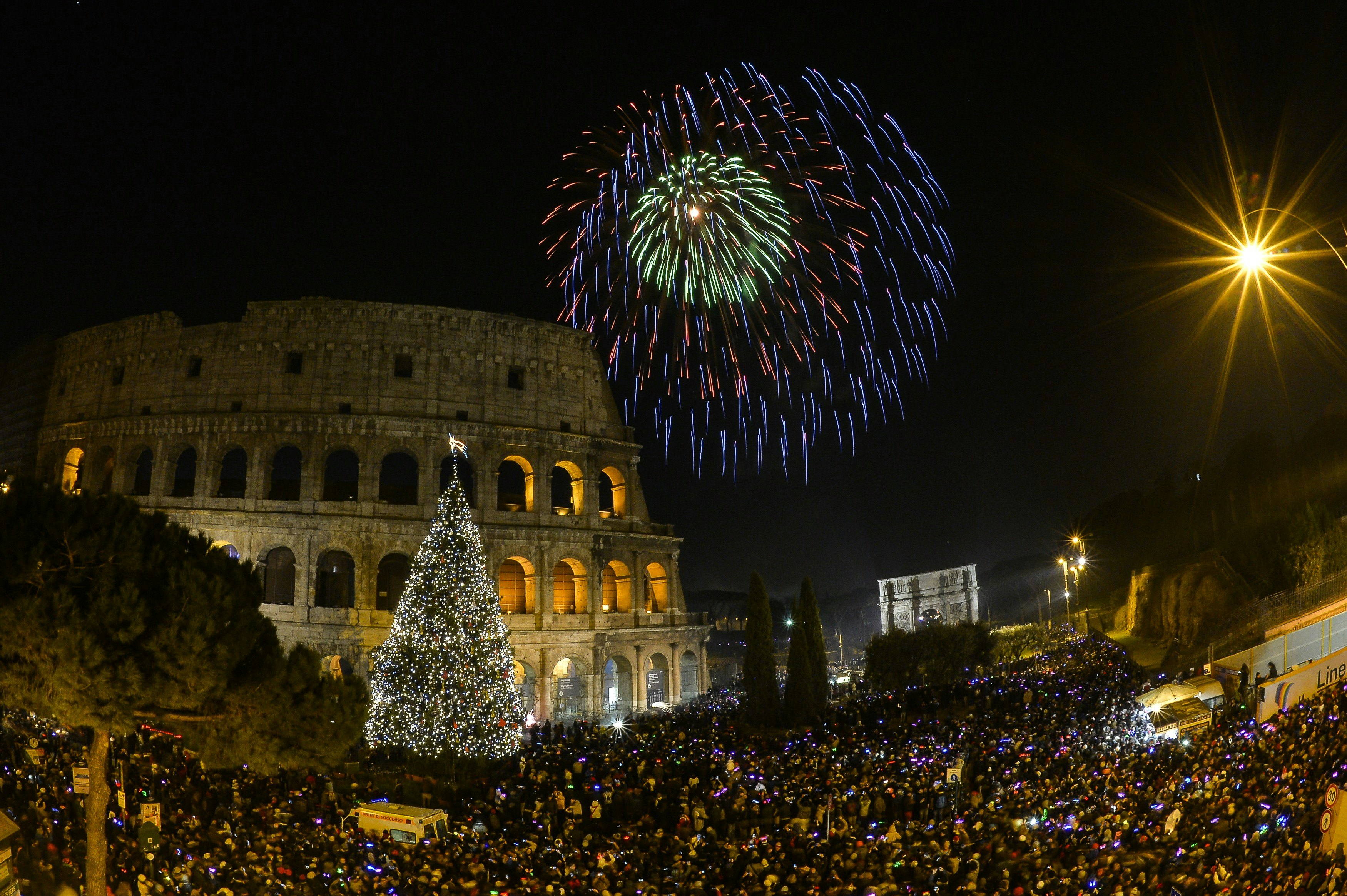 Crowds gather near a large round ancient amphitheater as fireworks light up the sky above