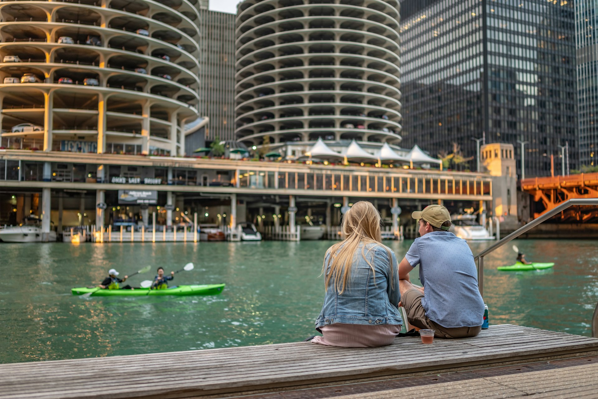 Two friends sit on a boardwalk that runs alongside a river in a city with tall buildings