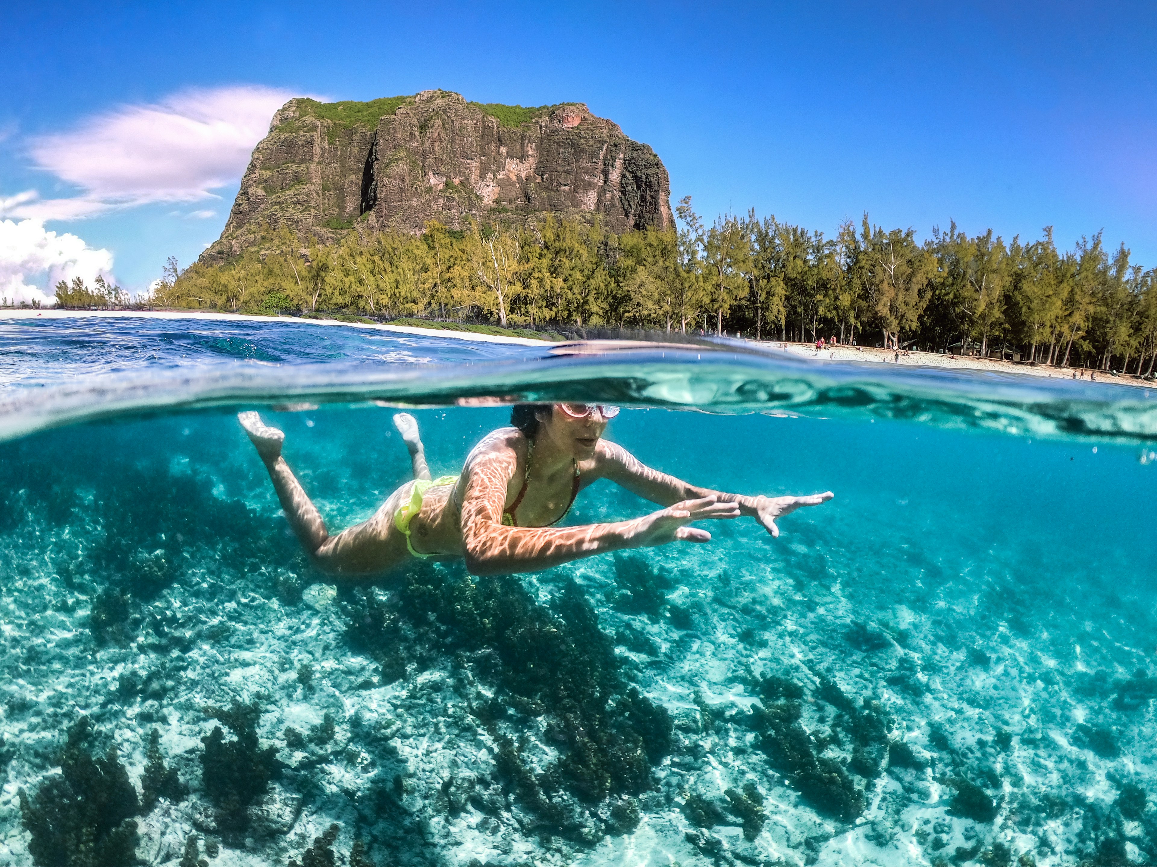 A woman snorkels through turquoise waters with a rocky outcrop on the beach in the background