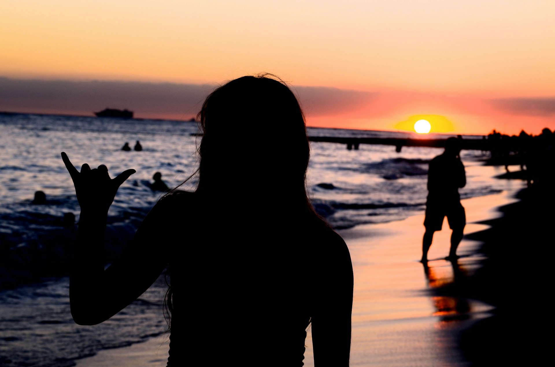 A female in silhouette as the sun sets over the beach holds up her hand in a hang loose sign or "shaka"