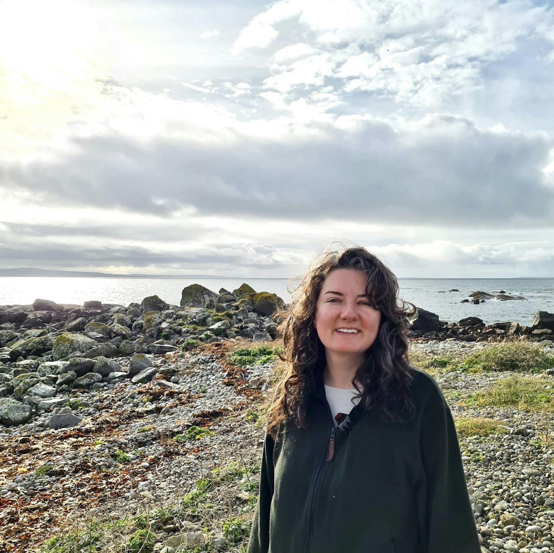 A woman wearing green stands on a rocky coastline. 