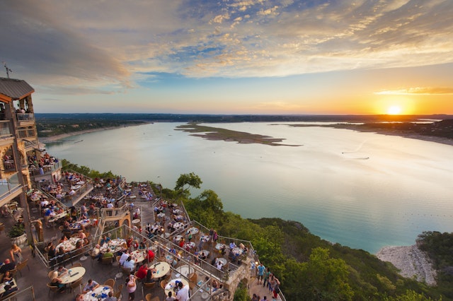 Diners eat on a terrace by a lake at sunset