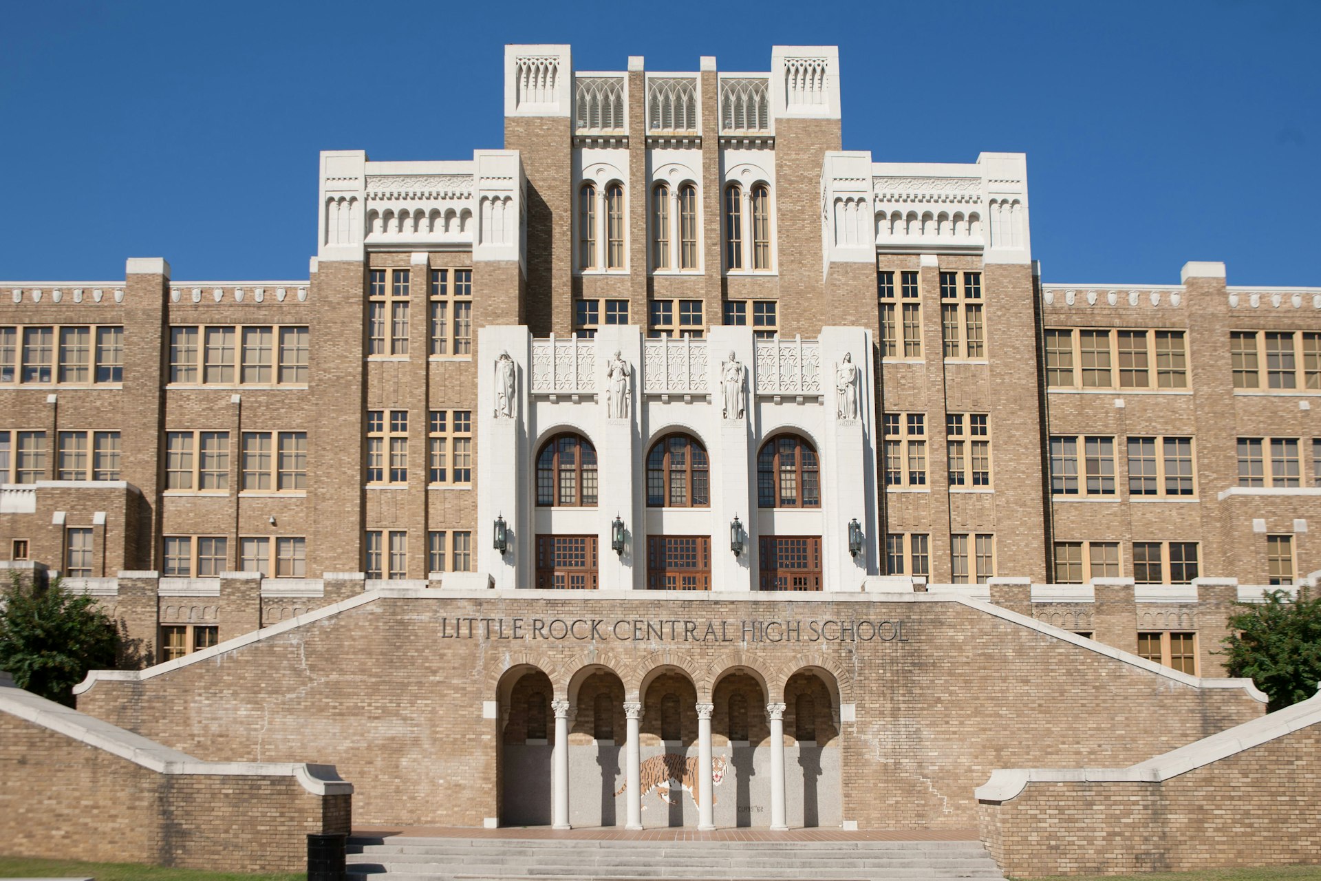 The brick exterior of Little Rock Central High School, with a row of four large statues above the doors and a mural of a tiger beneath the steps 