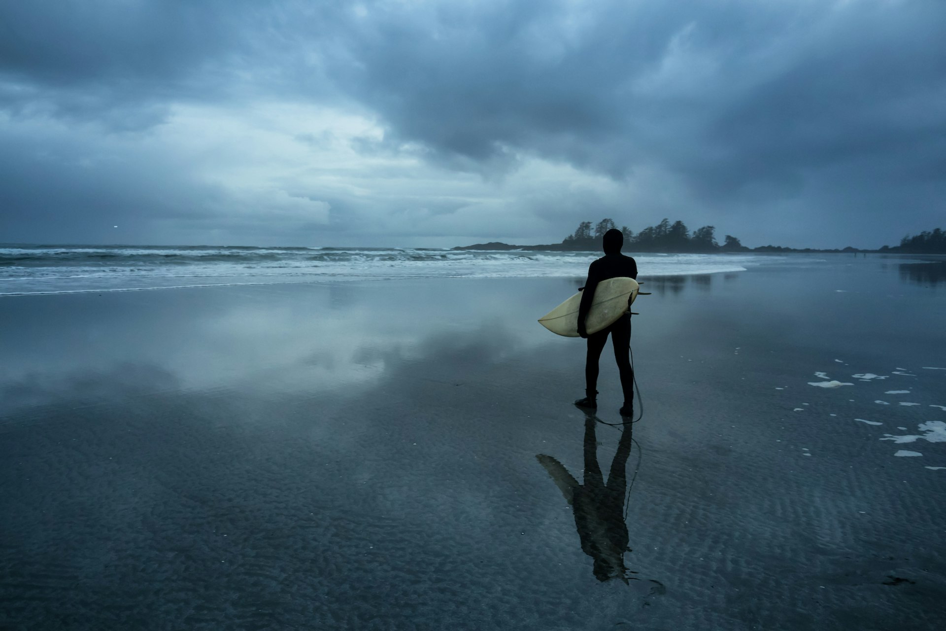 Surfer heading out in the ocean during a cloudy winter sunset, Chesterman Beach, Tofino, Vancouver Island, British Columbia, Canada