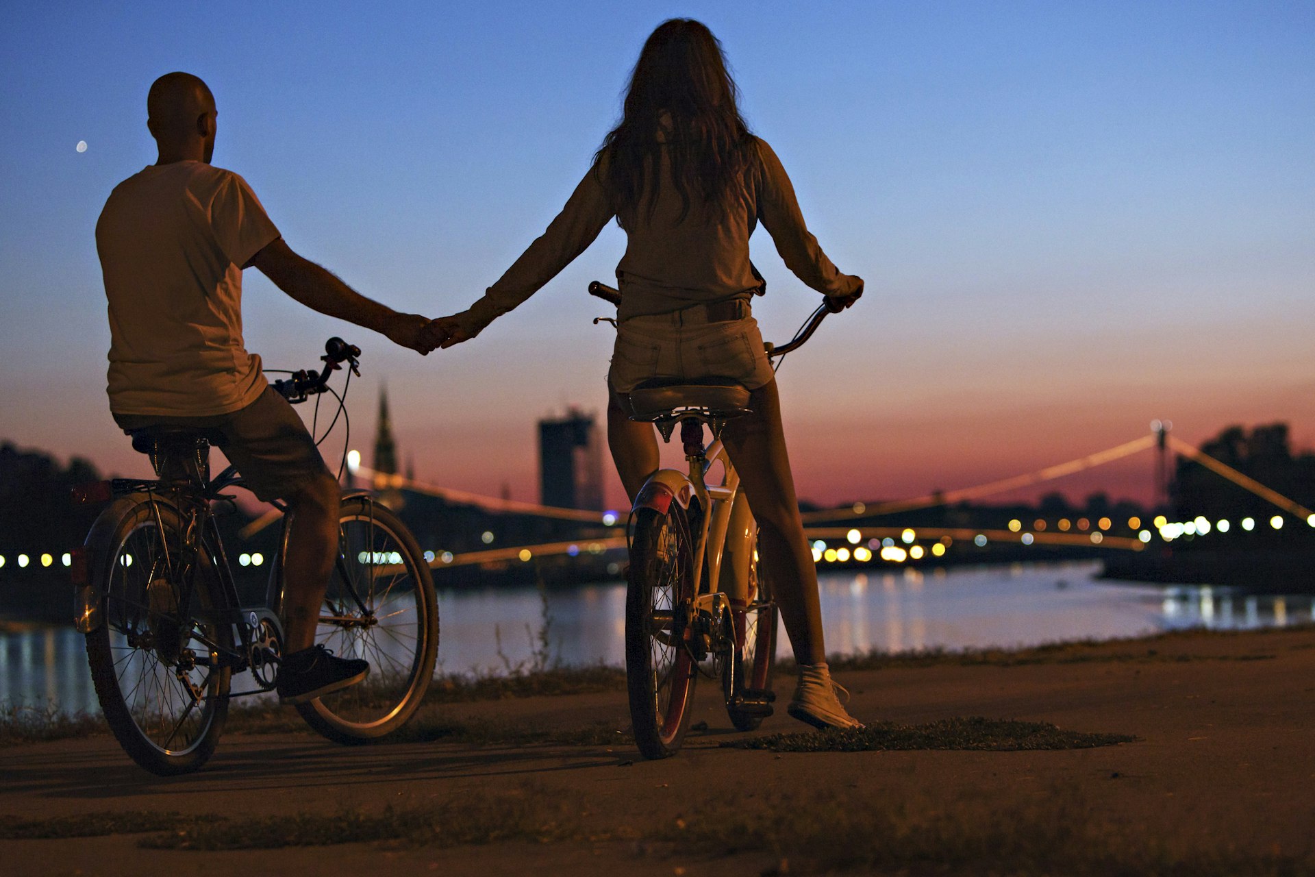 A young couple on bicycles, a boy and a girl, hold hands whilst looking across a bridge at night in Osijek, Croatia