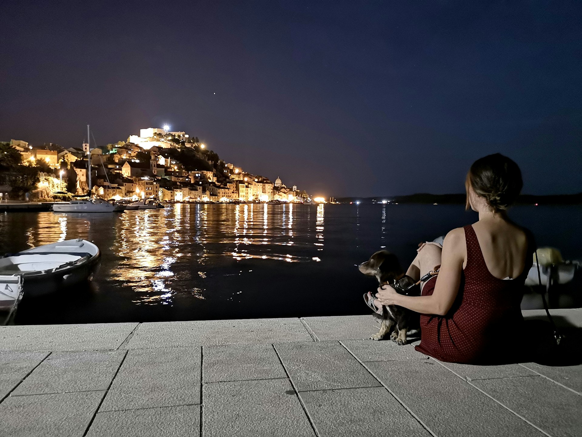 A woman with a dog looks out over the harbor at night at the illuminated town of Šibenik, Croatia
