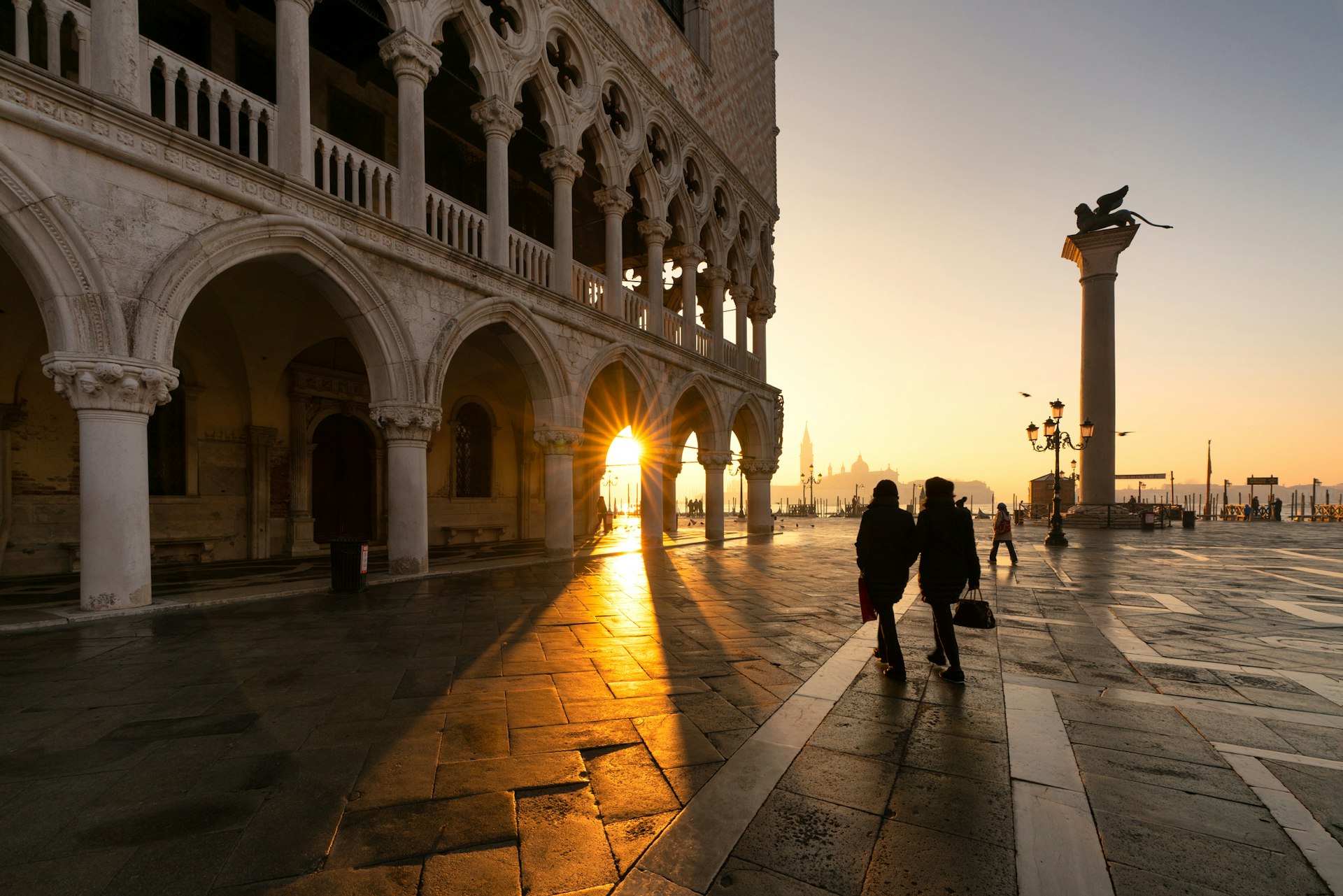 A couple walk near the empty arches of the Doge's Palace in Venice. Silhouetted by the morning sun breaking over the Grand Canal, the pair walk towards the water and a tall statue, the yellow rays of the sun peeking past the pillar holding up the Palace.