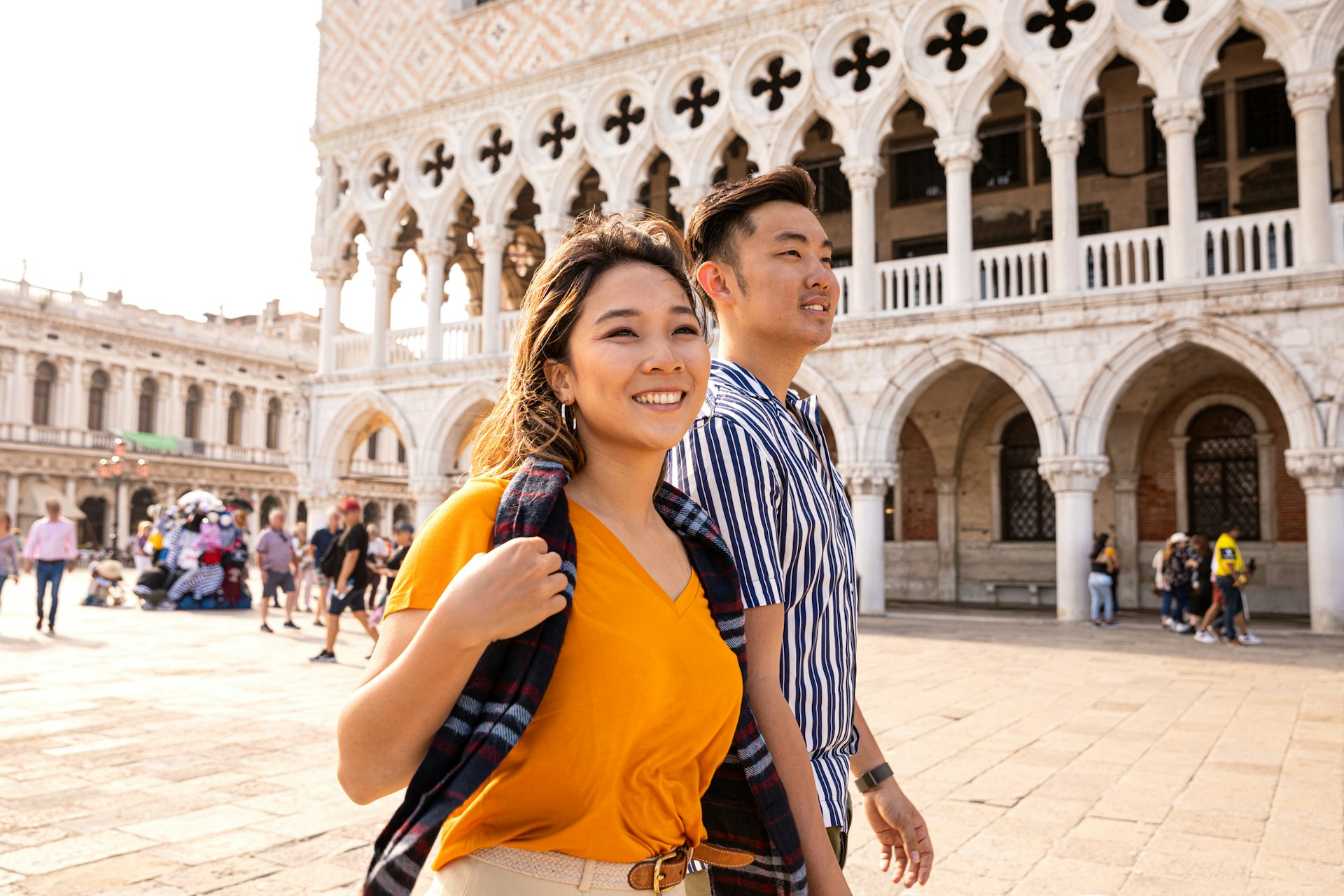 An Asian couple wander near Piazza San Marco in Venice. She is wearing a mustard yellow blouse with a burgundy scarf and he is in a white and blue striped shirt. Both are smiling as they look around. The Doge's Palace is visible in the background alongside a tourist stall and some blurred walkers.