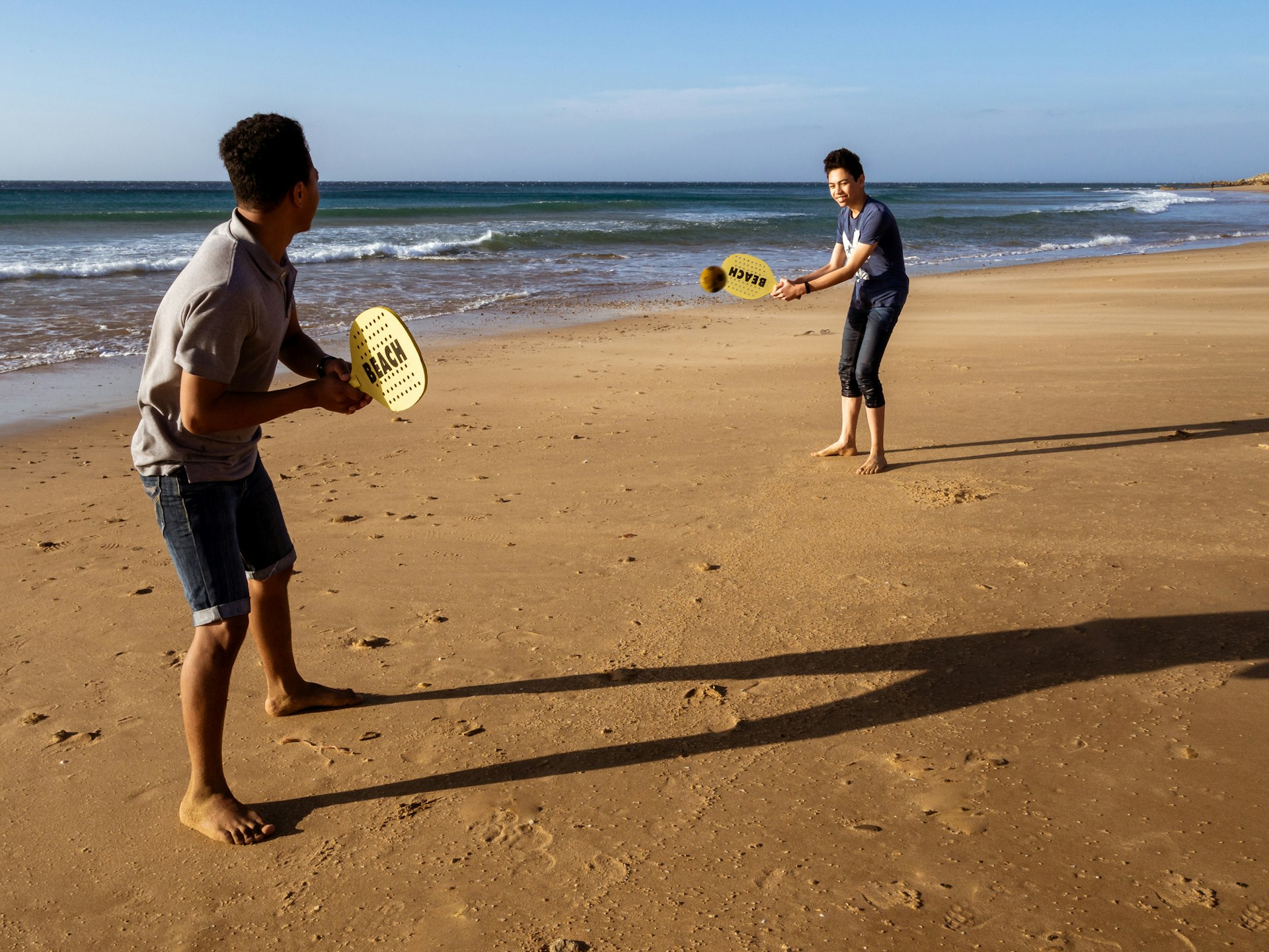 Two smiling teens play racket ball on the orange sand of Achakar beach in Tangier, Morocco