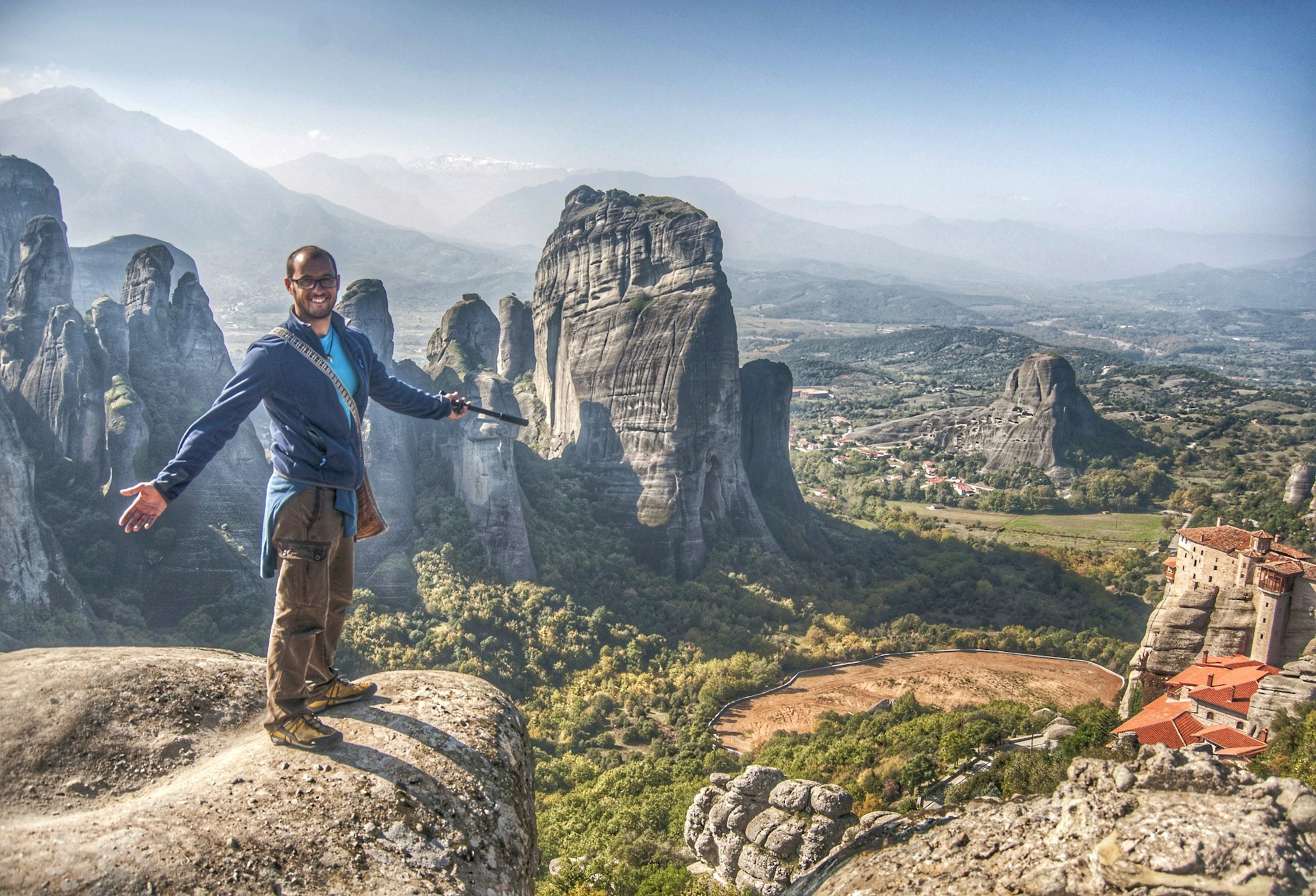 A man stands on a rocky outcrop, clearly delighted at the view that stretches into the distance with peaks topped with monasteries
