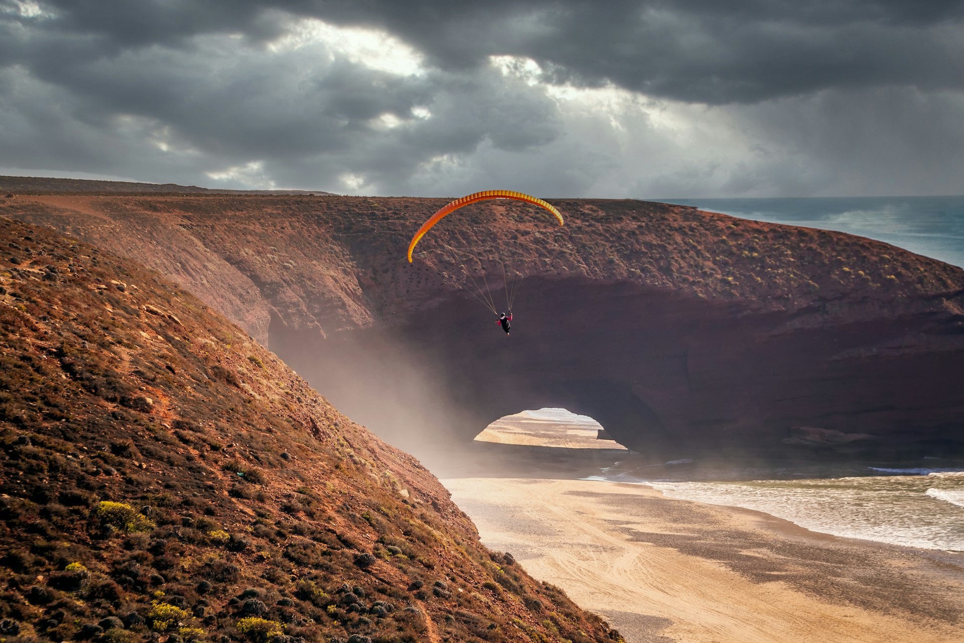 A paraglider drops down onto the red sand next to the land arch of Legzira beach, in Morocco at sunset as wind whips up the grains of sand
