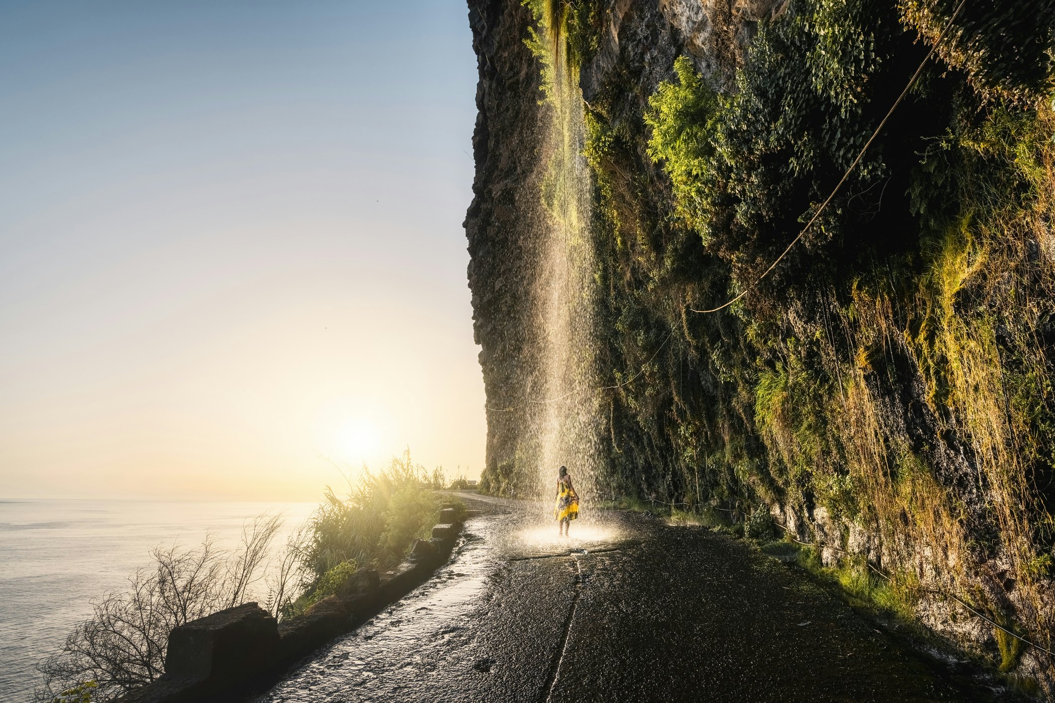 A woman stands under a waterfall which pours off a cliff in the middle of the road in Madeira with the sea out to the left