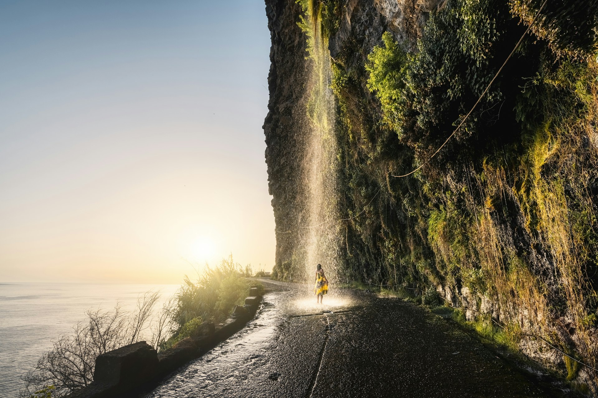 A woman stands under a waterfall which pours off a cliff in the middle of the road in Madeira with the sea out to the left
