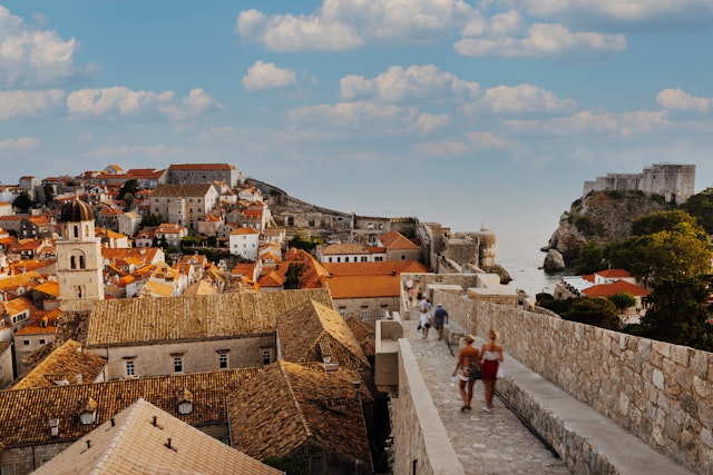 People walk along the old city walls that surround the copper-colored roofs of an old town by the sea.