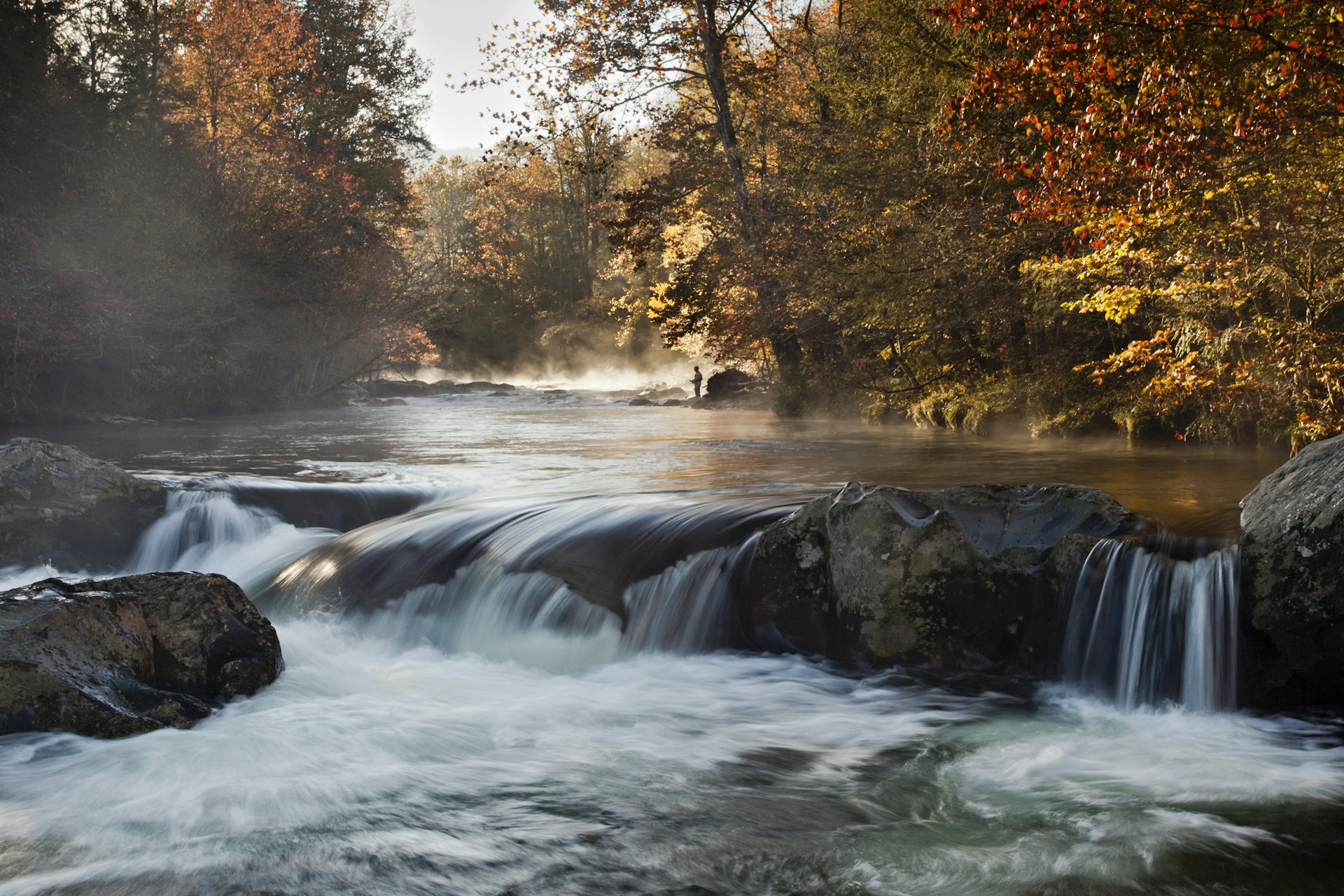 A fisher stands in the background of an autumn scene with mist still floating on the river and water flowing over rocks in the foreground