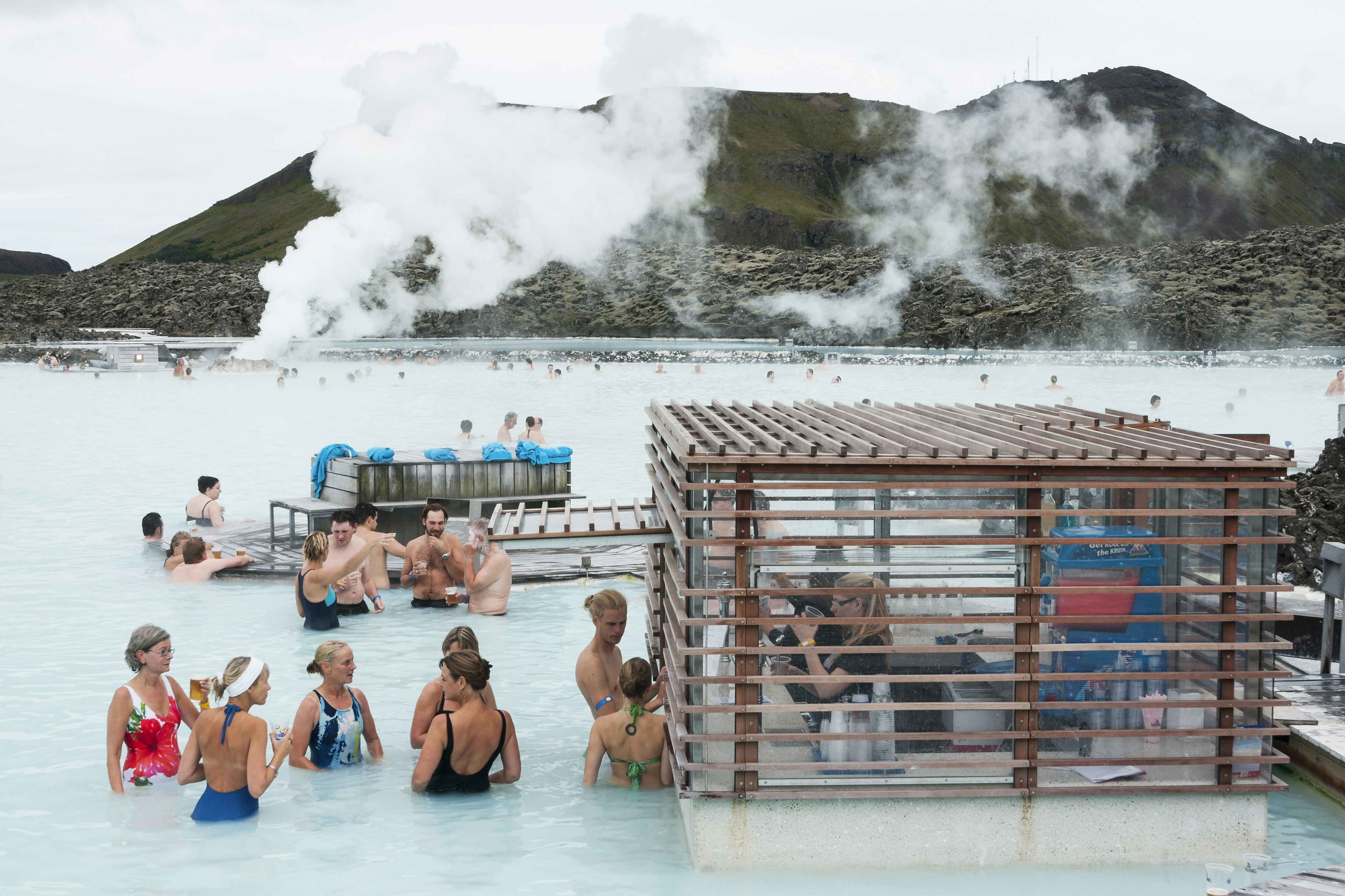 People in bathing suits gather in a geothermally heated pool. Some order drinks from an adjacent bar. Steam from geothermal vents rises in the distance.