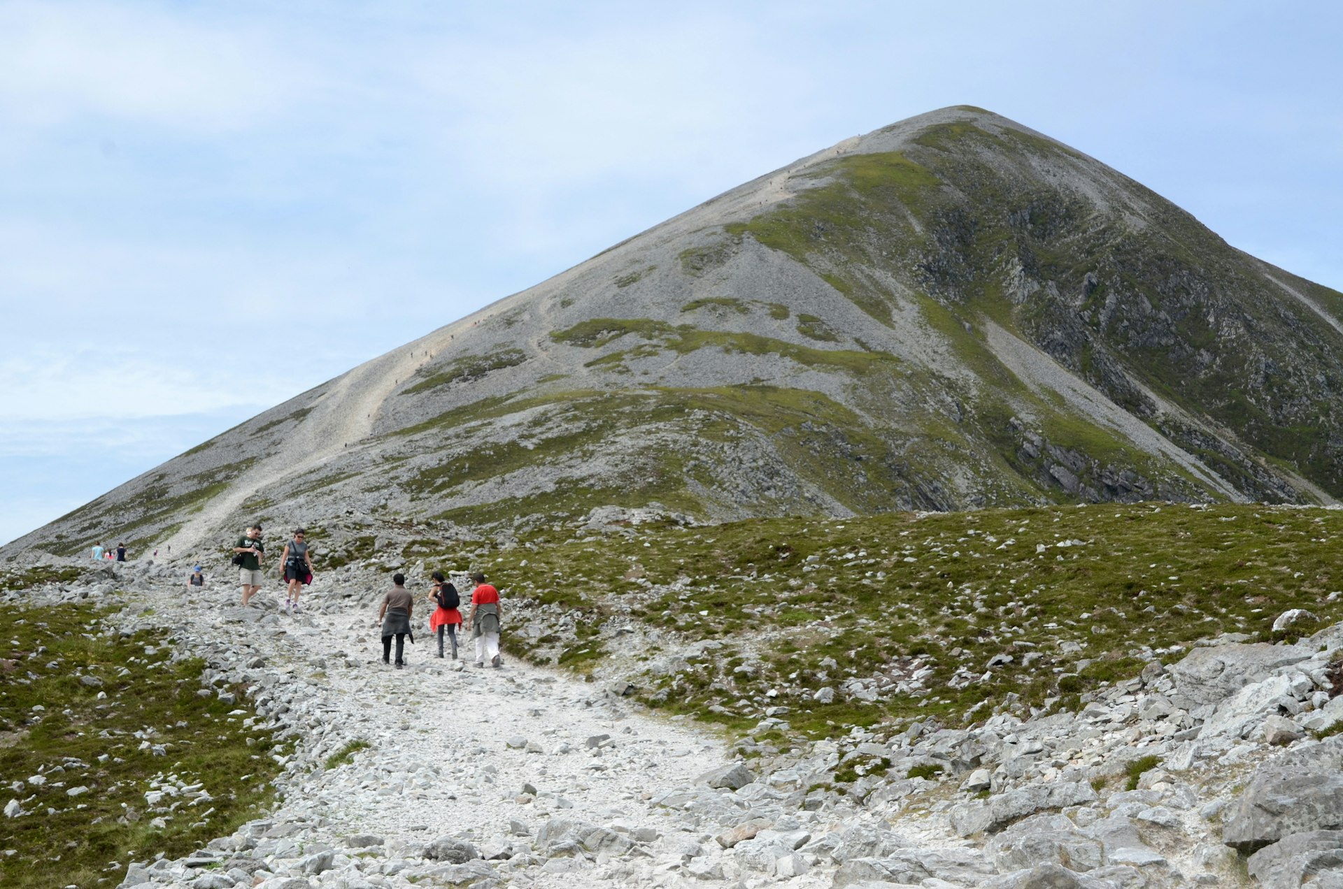 Hikers ascend and descend a steep gravel path leading towards a mountain peak