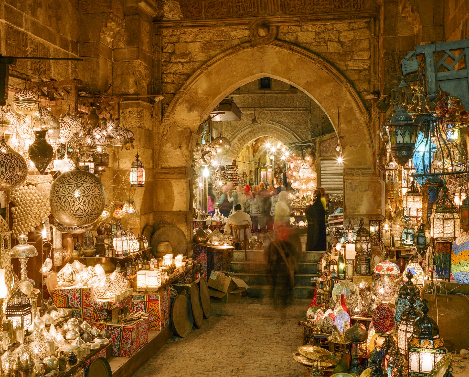 People walk down alleyways lit with intricate lamps in a busy marketplace