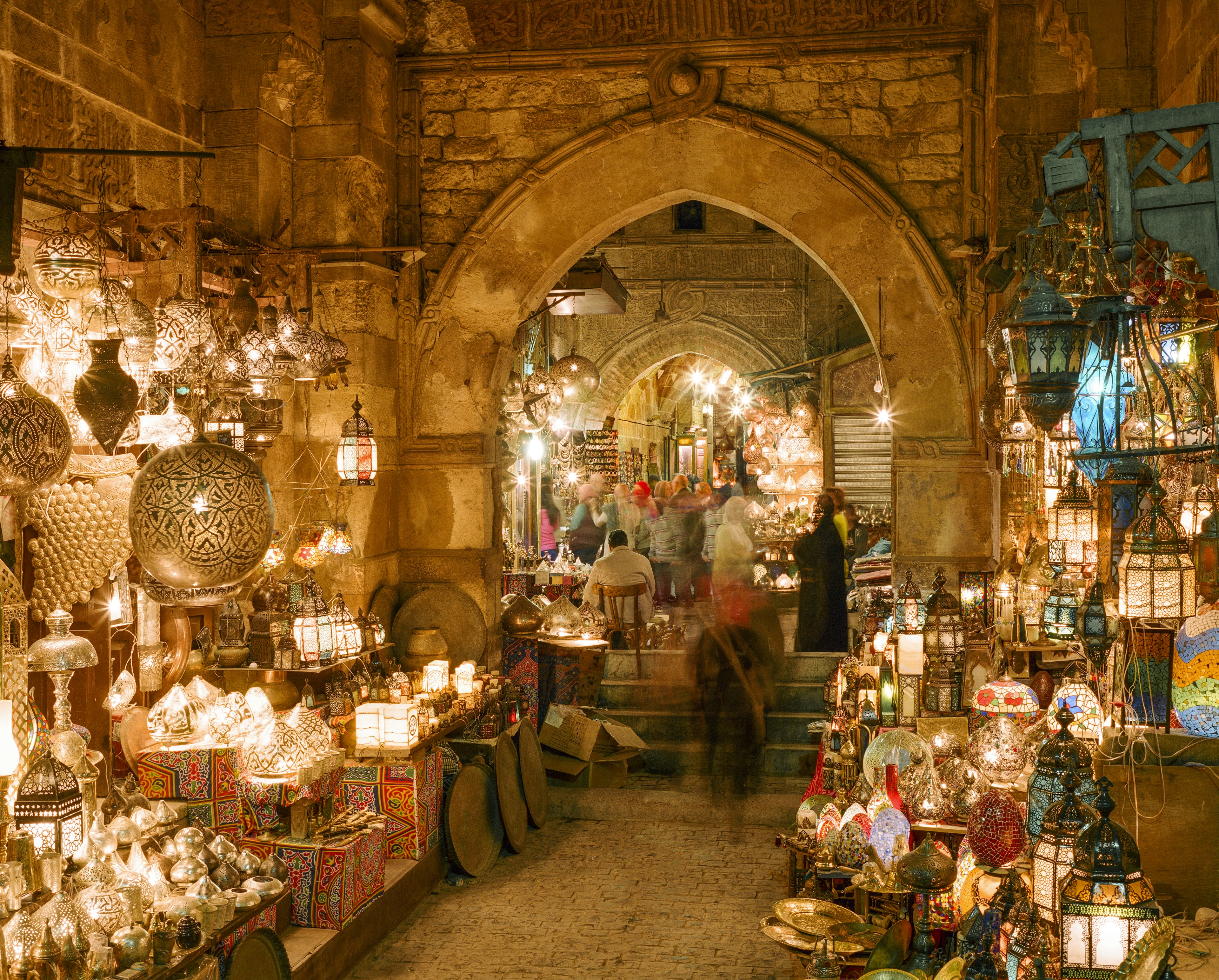 People walk down alleyways lit with intricate lamps in a busy marketplace