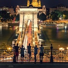 Five people looking out over Széchenyi Chain Bridge in Budapest, Hungary, at night.