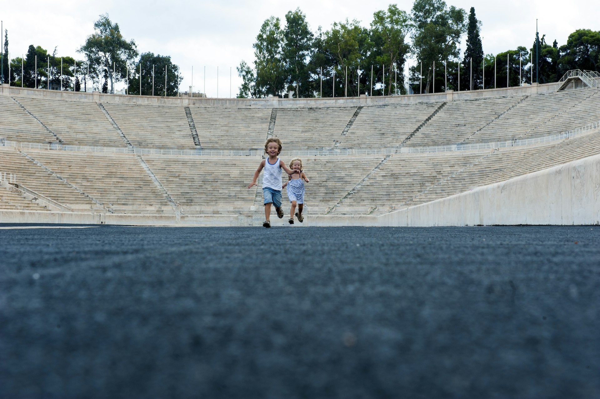 Two young children, a boy and a girl, run along an open area within a stadium