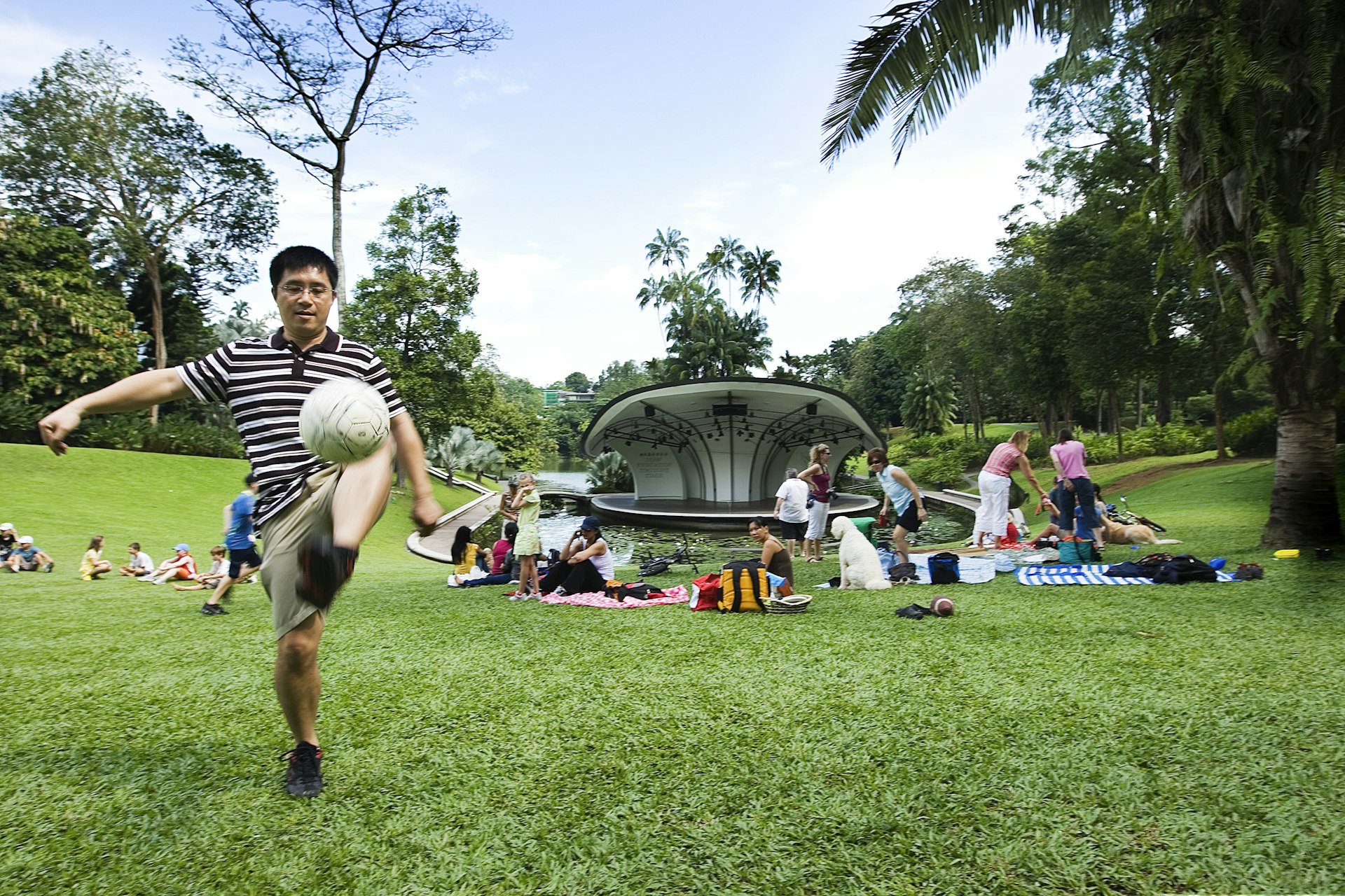 A man kicks a football while people picnic in the background at Singapore Botanic Gardens