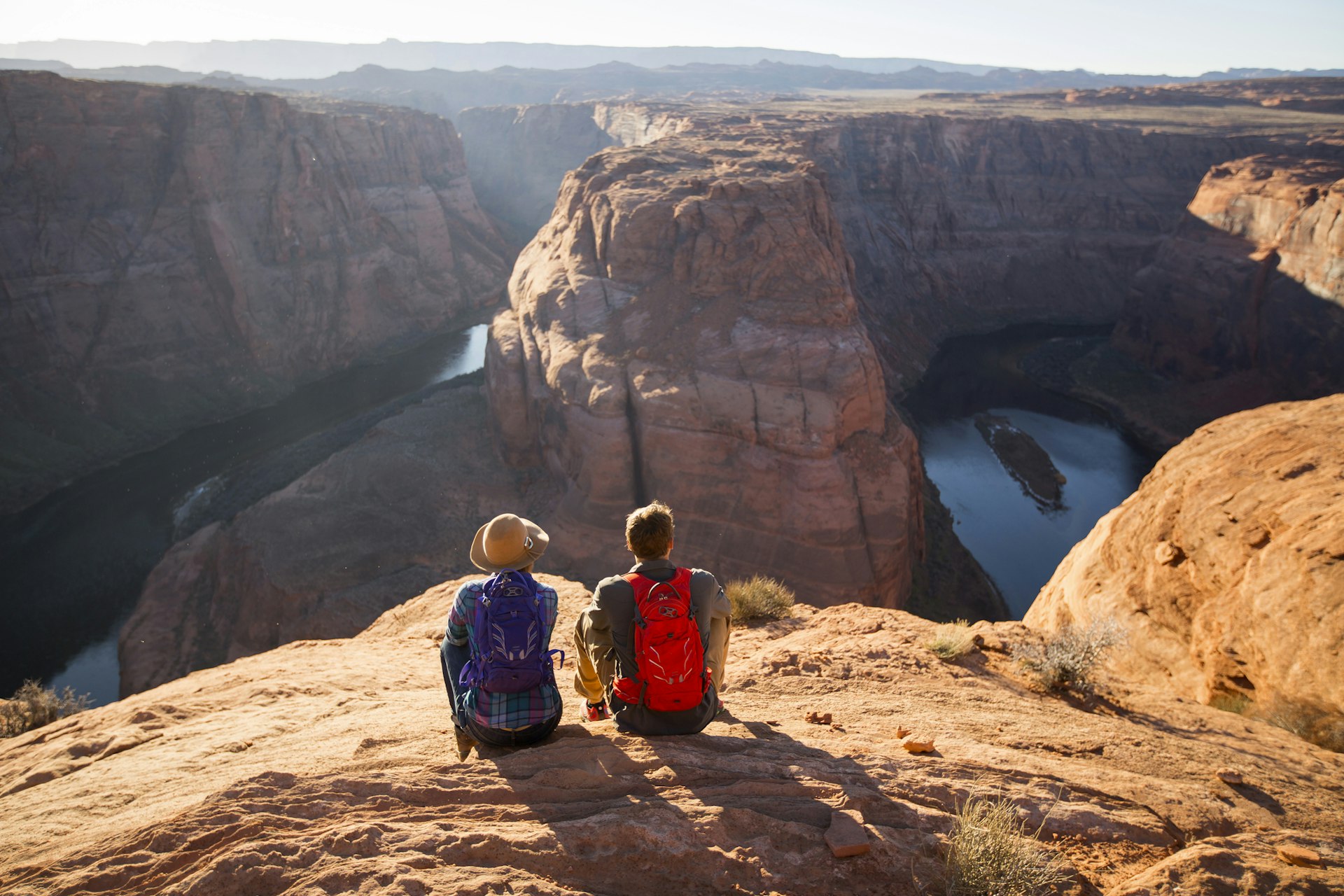 Two people sit on the cliff above a vast curve in a river surrounded by red rock formations