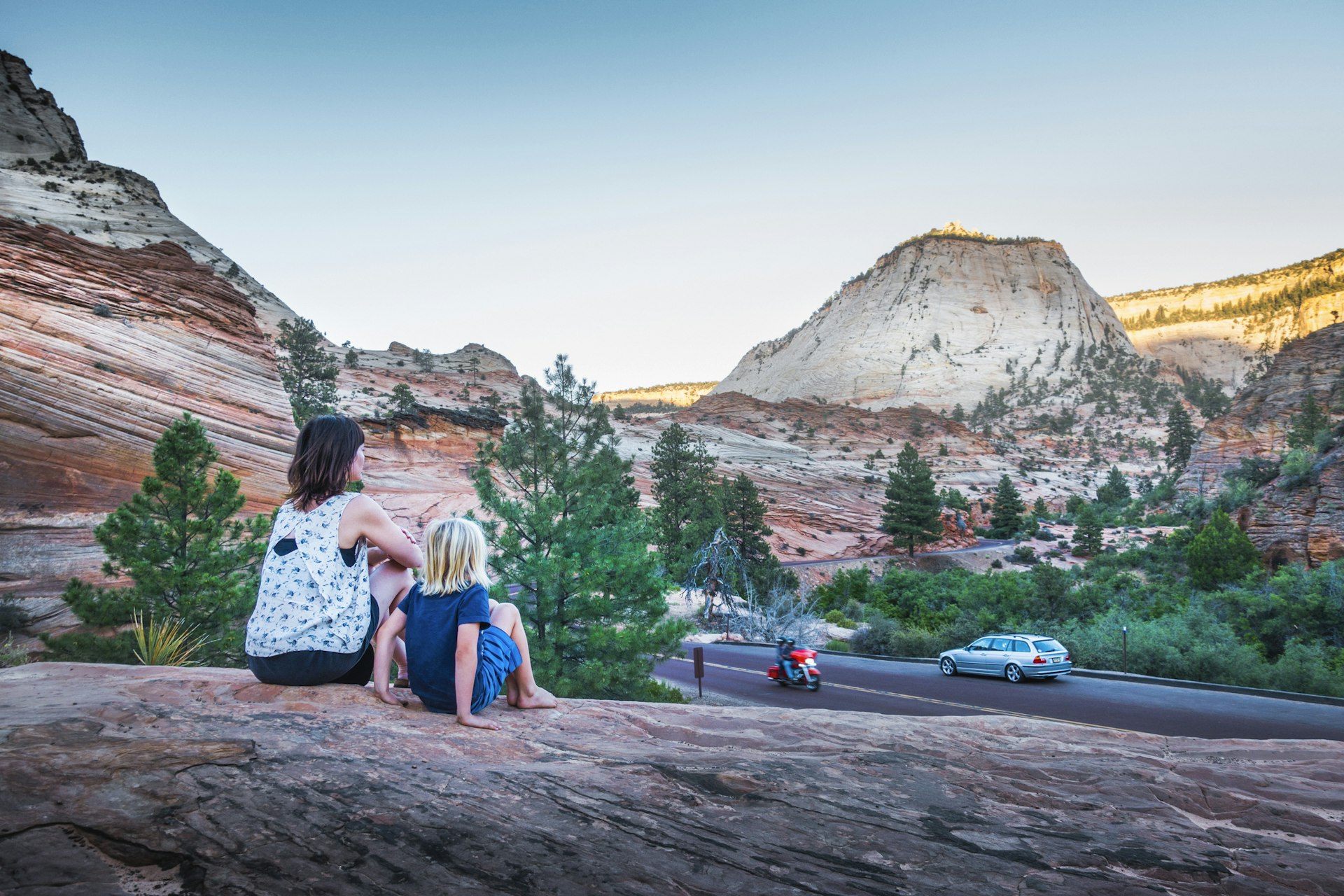 A woman and a child sit on the rocks above a red rock landscape as a car and motorbike drive by