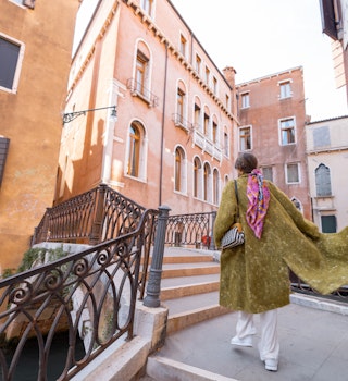 Young woman walking beautiful streets at water canals in Venice. Concept of happy vacations in Italy. Caucasian stylish woman in coat with colorful shawl Young woman walking beautiful streets at water canals in Venice. Concept of happy vacations in Italy. Caucasian stylish woman in coat with colorful shawl