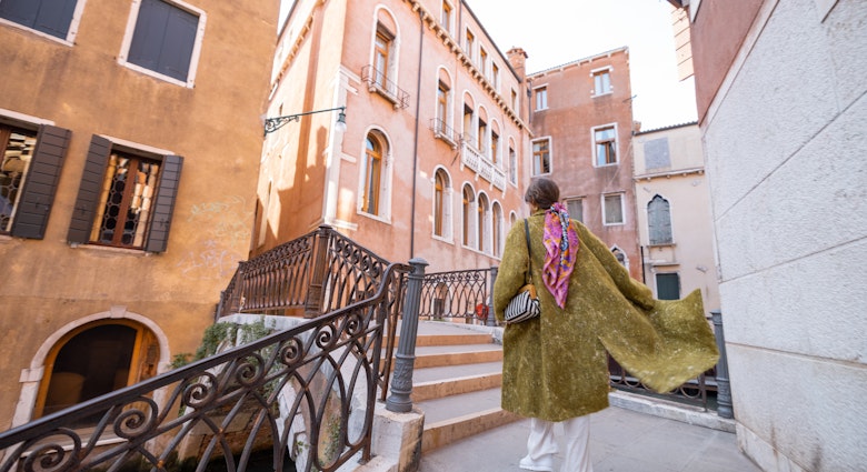 Young woman walking beautiful streets at water canals in Venice. Concept of happy vacations in Italy. Caucasian stylish woman in coat with colorful shawl Young woman walking beautiful streets at water canals in Venice. Concept of happy vacations in Italy. Caucasian stylish woman in coat with colorful shawl