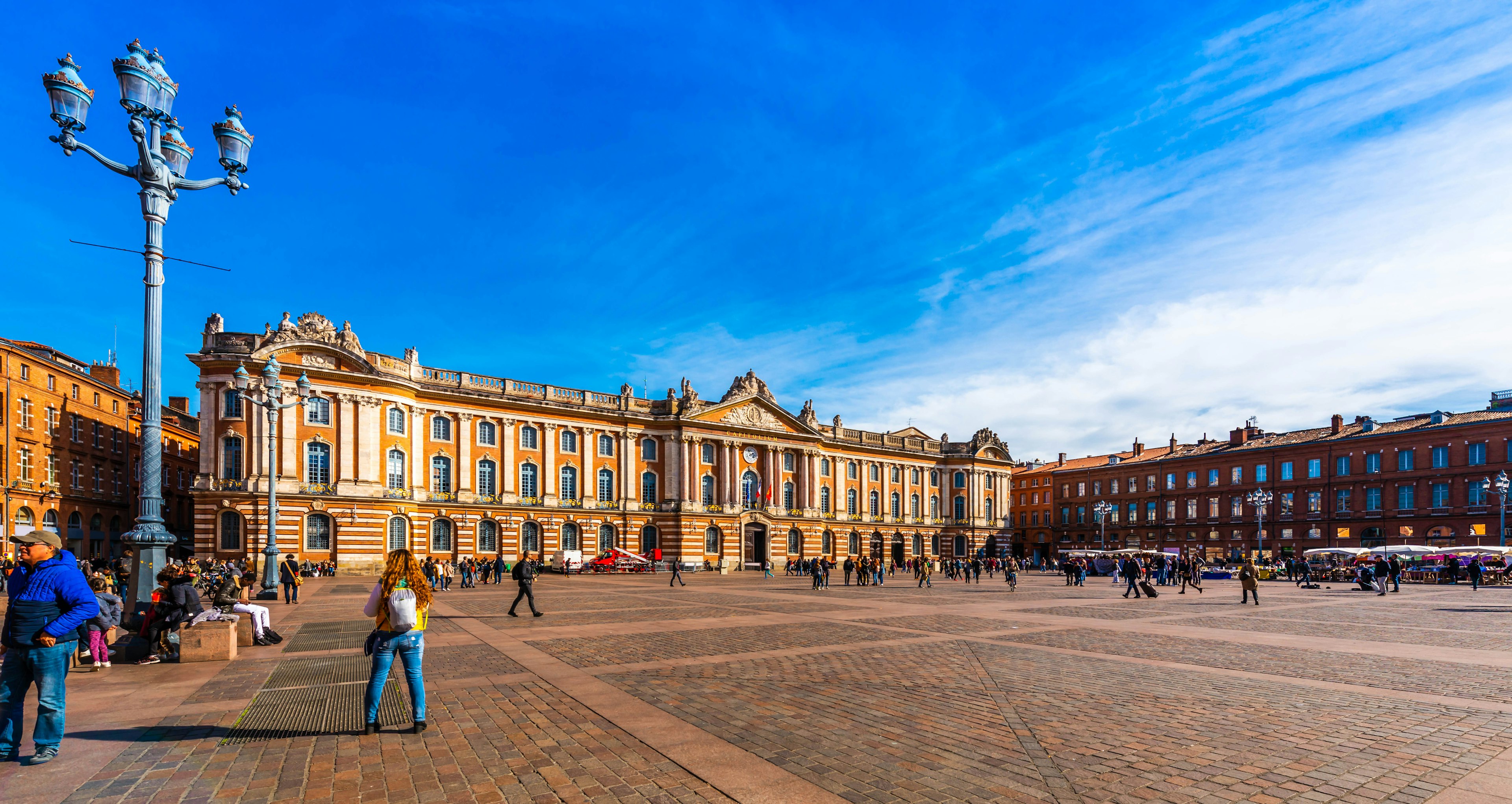 A large city square busy with people, with one very long building dominating one side of it