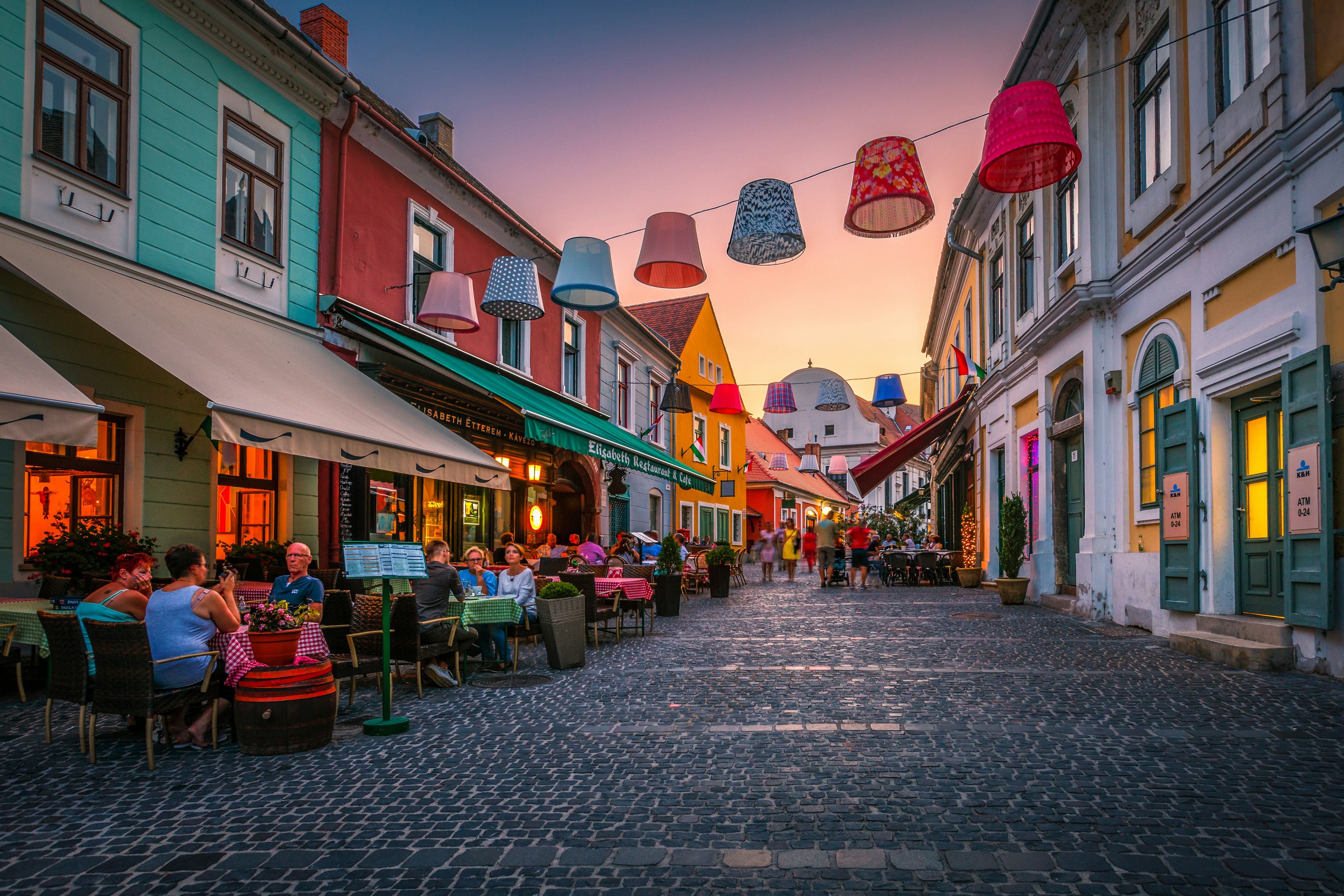 People seated at restaurants and coffee shops in the old town of Szentendre during sunset.