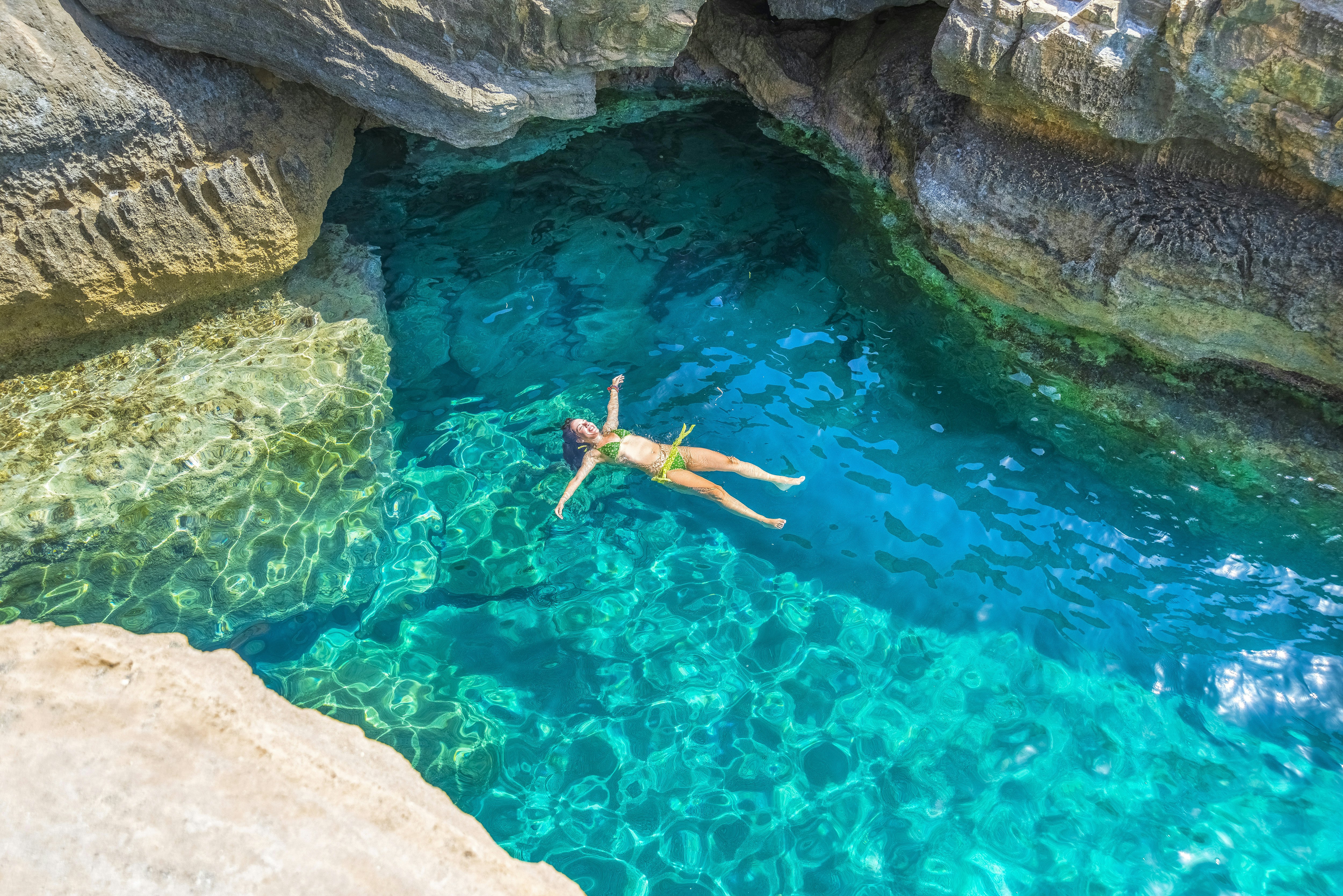 A woman smiles as she floats in a turquoise ocean in a bay sheltered by rocks