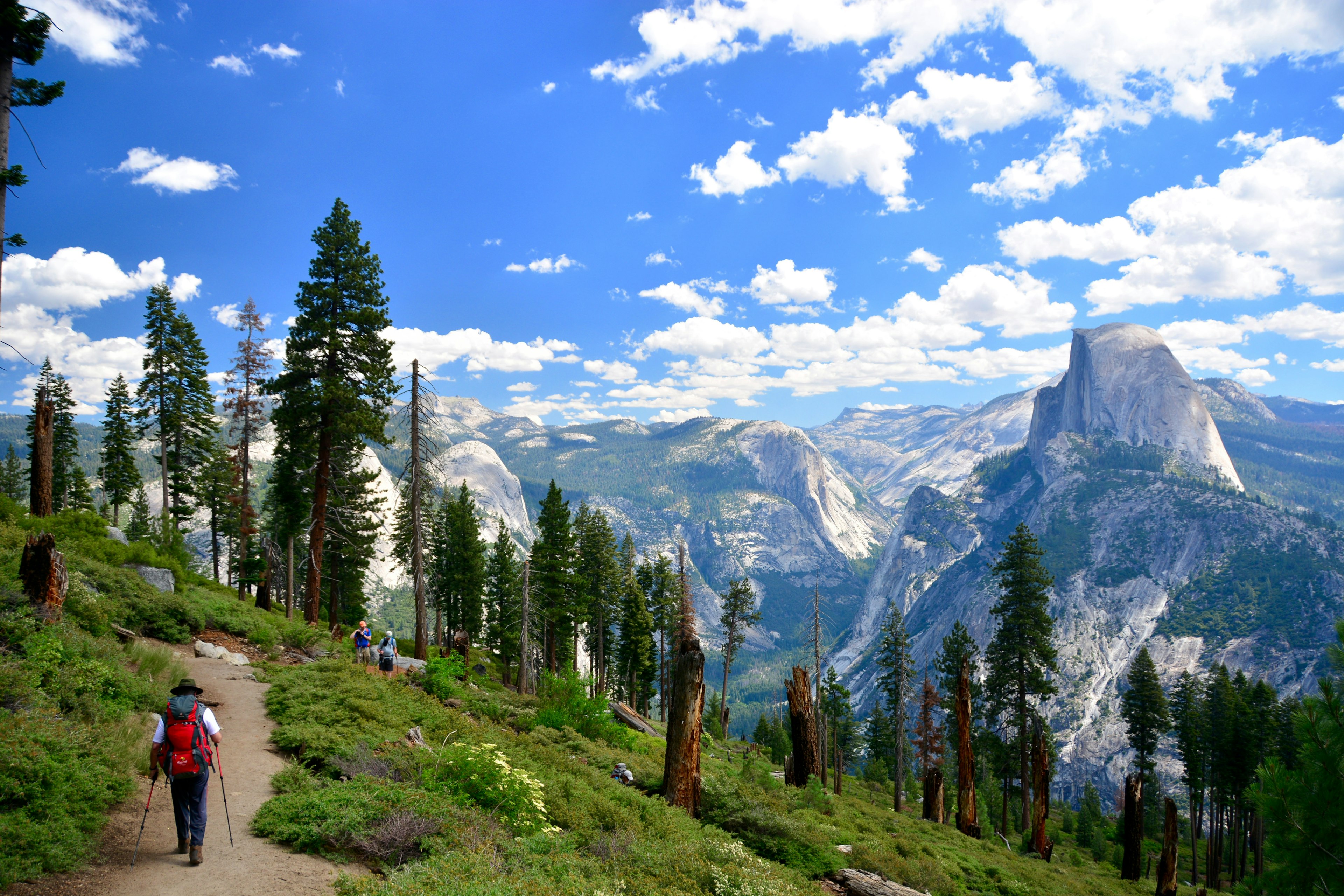 A hiker follows a dirt path on a sunny day with dramatic rock formations and tall cliffs to the left