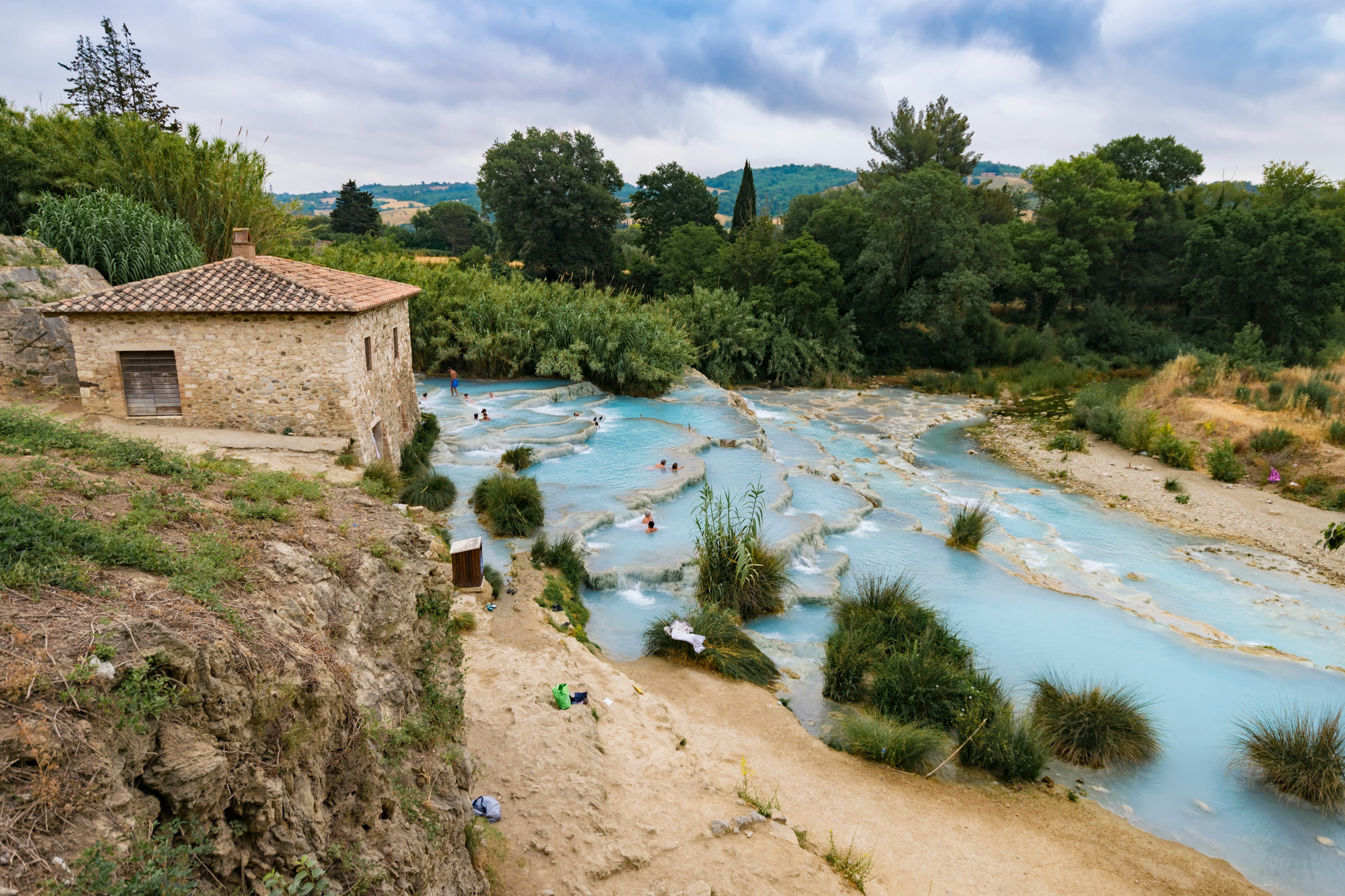 People bathing at a natural spa with waterfalls in Saturnia with a stone building in the foreground.