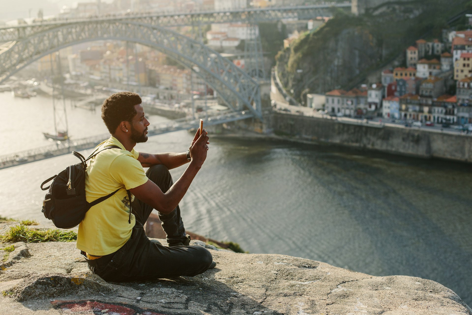 A man sits at a viewpoint of a city over a river and takes a photograph on his phone