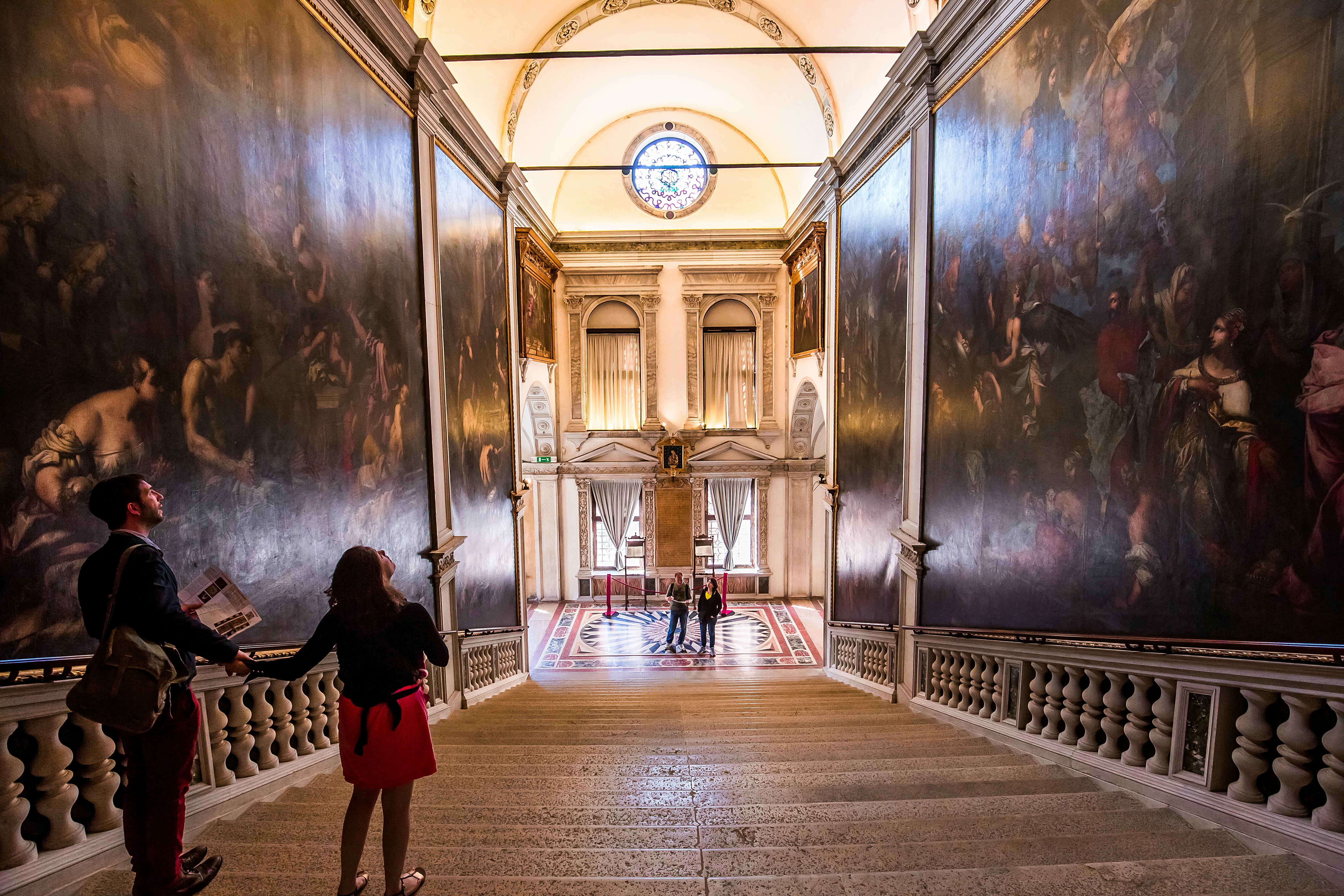 A man and woman both stand on a set of stone stairs admiring the intricate murals painted on the surrounding walls