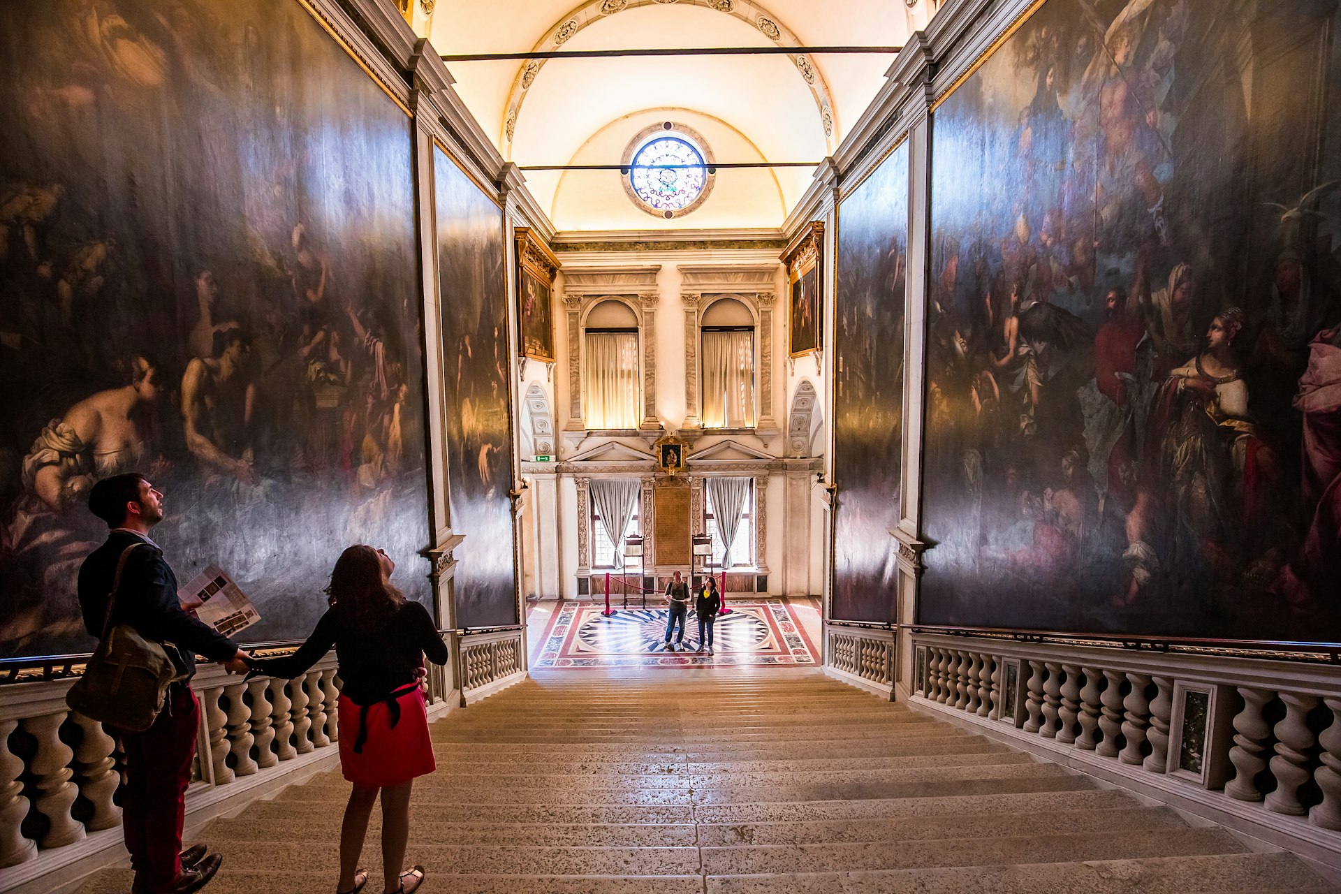 A man and woman both stand on a set of stone stairs admiring the intricate murals painted on the surrounding walls at Scuola Grande di San Rocco in Venice. 