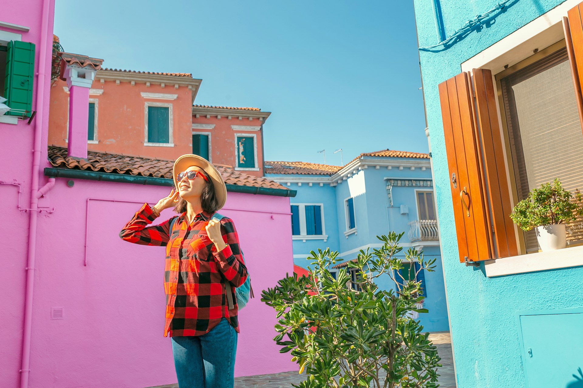 A happy South Asian woman in red sunglasses holds onto her hat in the sunshine as she wanders past the bright pink houses of Burano near Venice, Italy