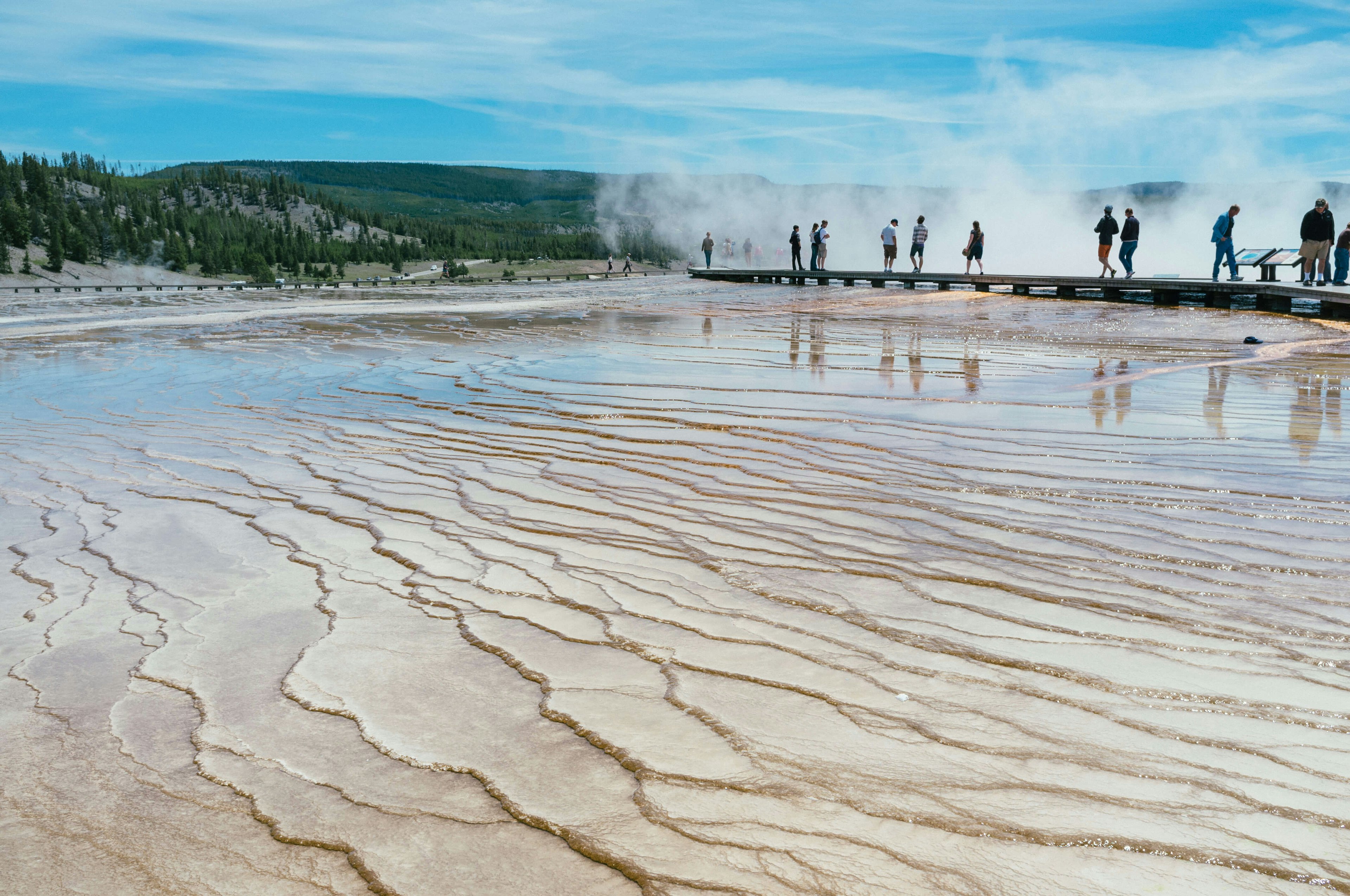 Tourists walking around the Grand Prismatic spring in Yellowstone National Park, low-angle view with mud and shallow water in the foreground