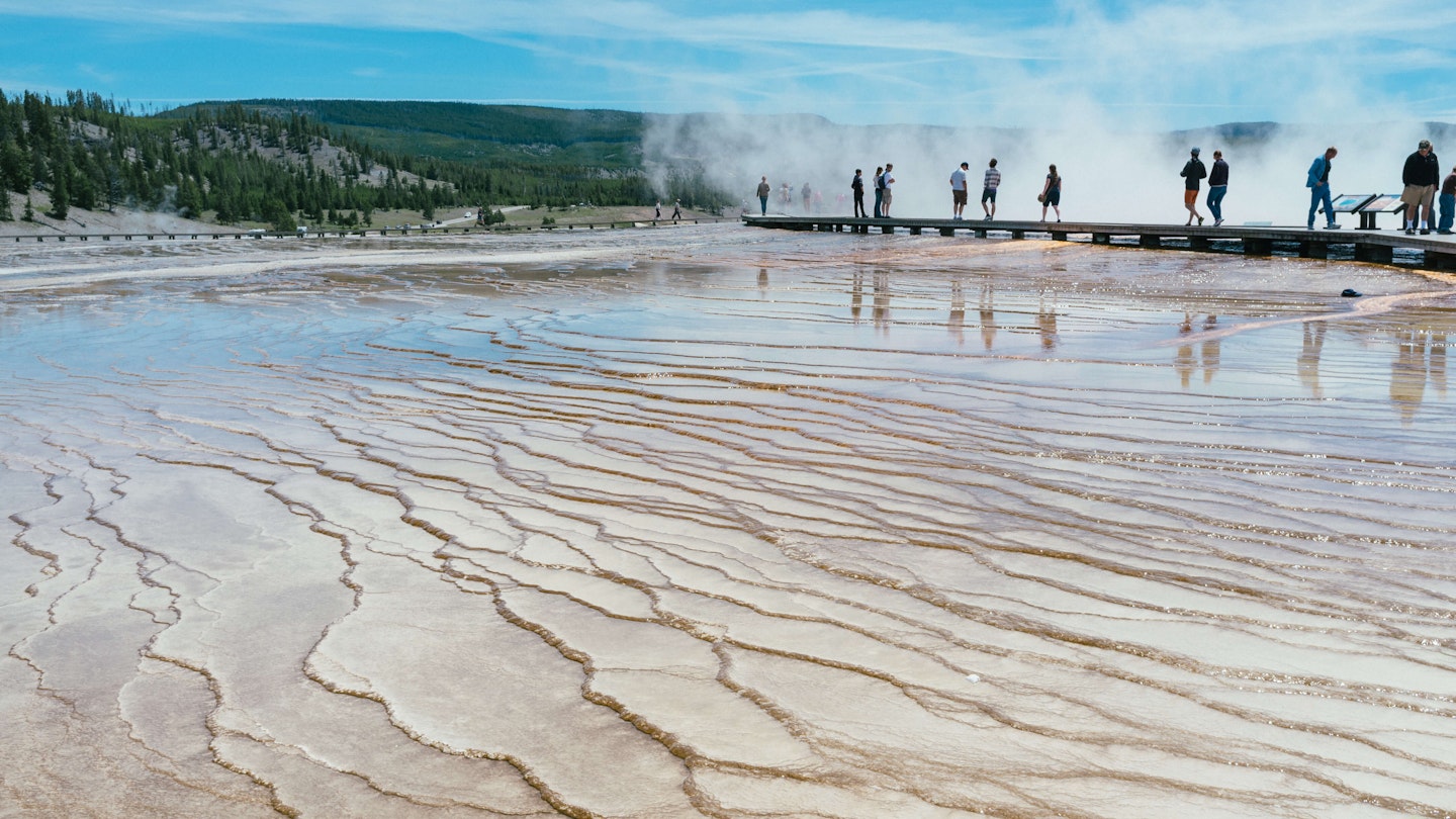 Tourists walking around the Grand Prismatic spring in Yellowstone National Park, low-angle view with mud and shallow water in the foreground