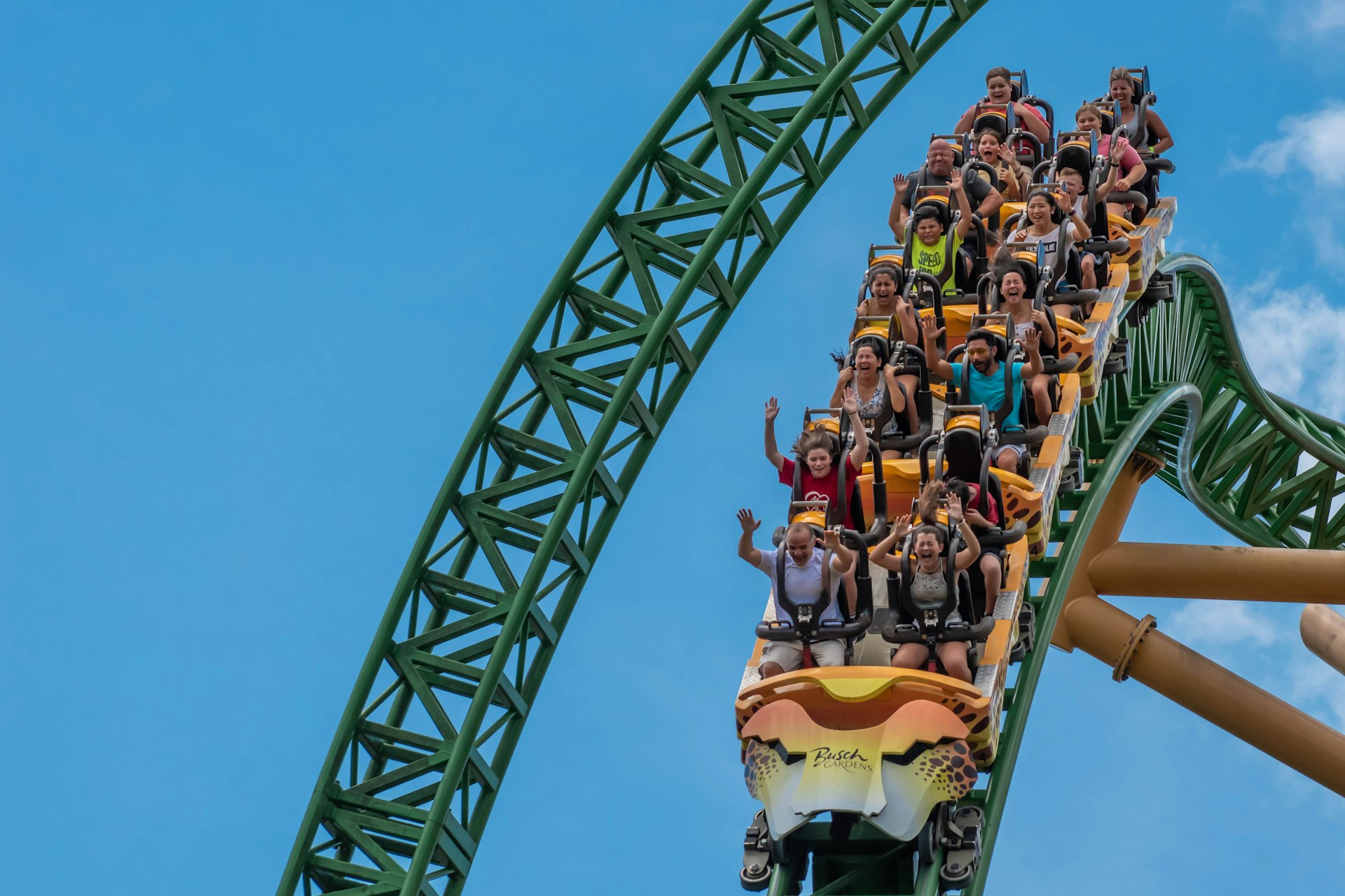 People on a rollercoaster with their arms up, screaming and smiling, against a light blue cloudy sky