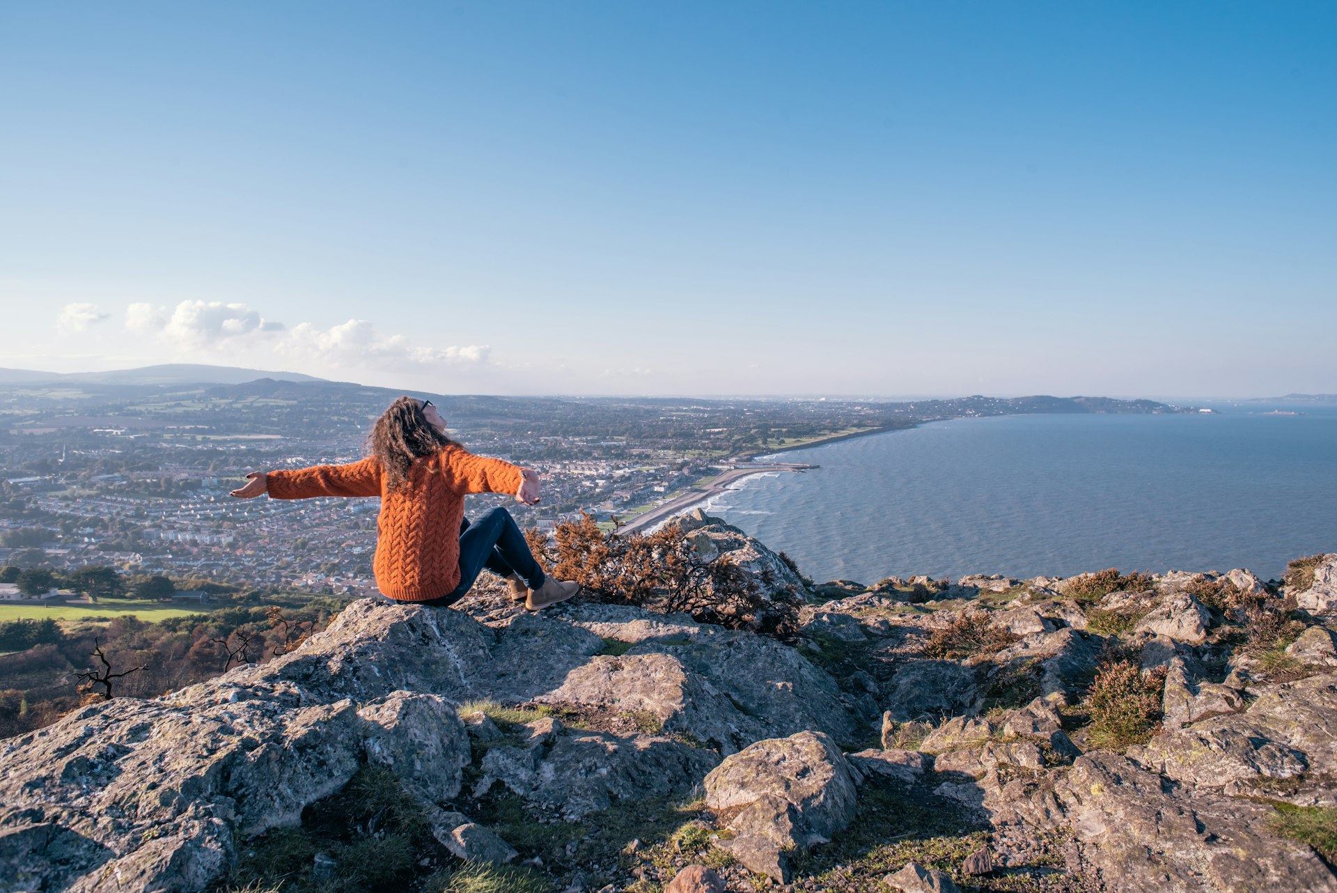 A woman sits on a rocky viewpoint above the coastline and throws her arms up in the air
