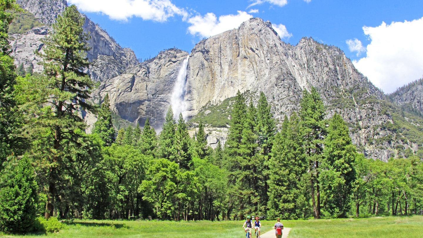 Yosemite National Park - The view on beautiful Yosemite Falls on the big rocky cliff surrounded by green trees, tourists walking and biking on pathway in the meadow with soft green grass. June 7, 2018