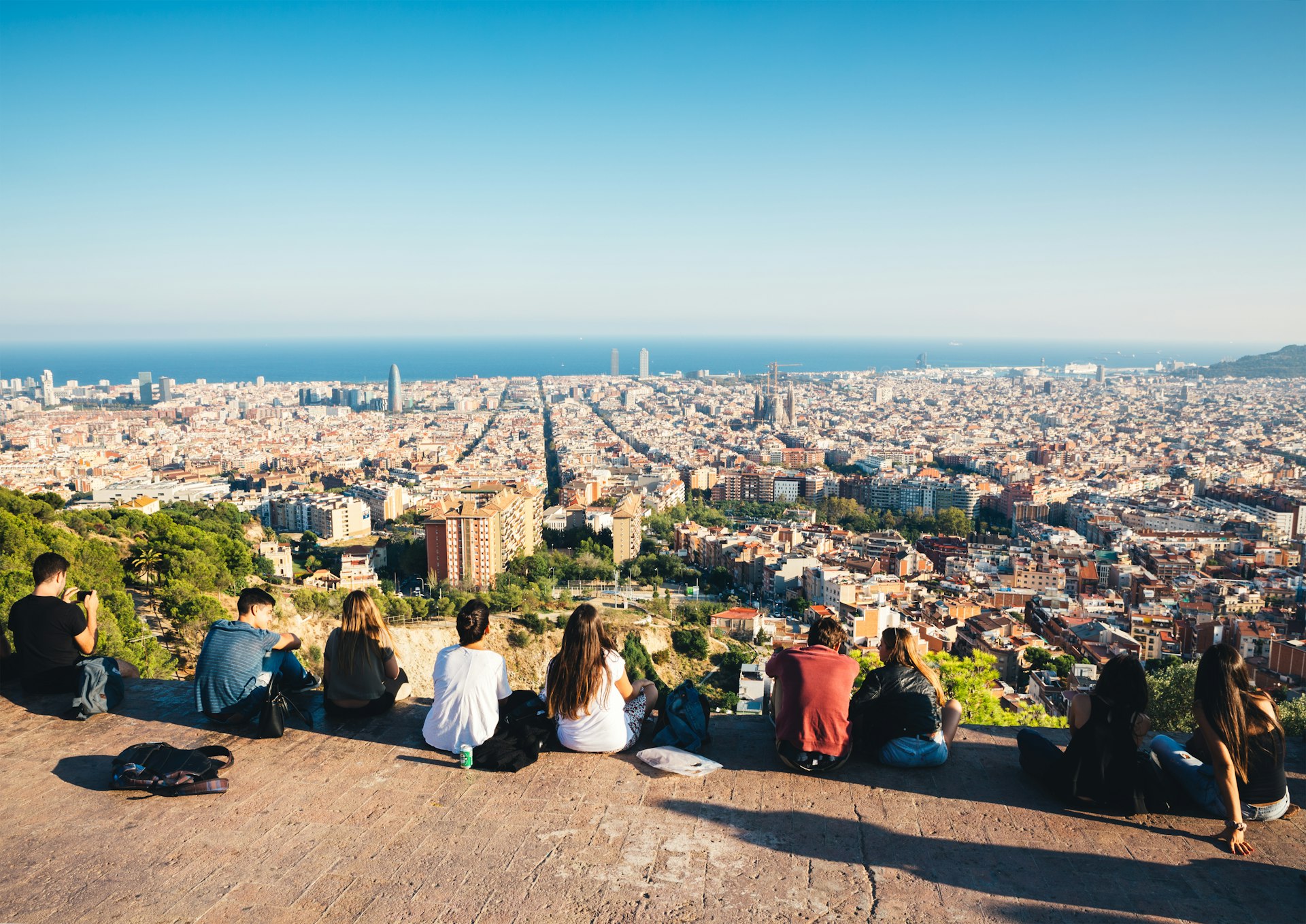 Group of young people are sitting on the wall watching Barcelona cityscape with Sagrada Familia and sea in the distance