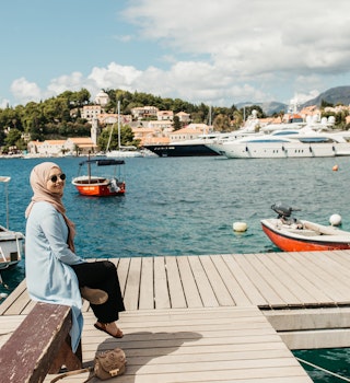 woman with hijab sitting on bench by the sea.  Female tourist exploring Cavtat. She is looking to the camera.