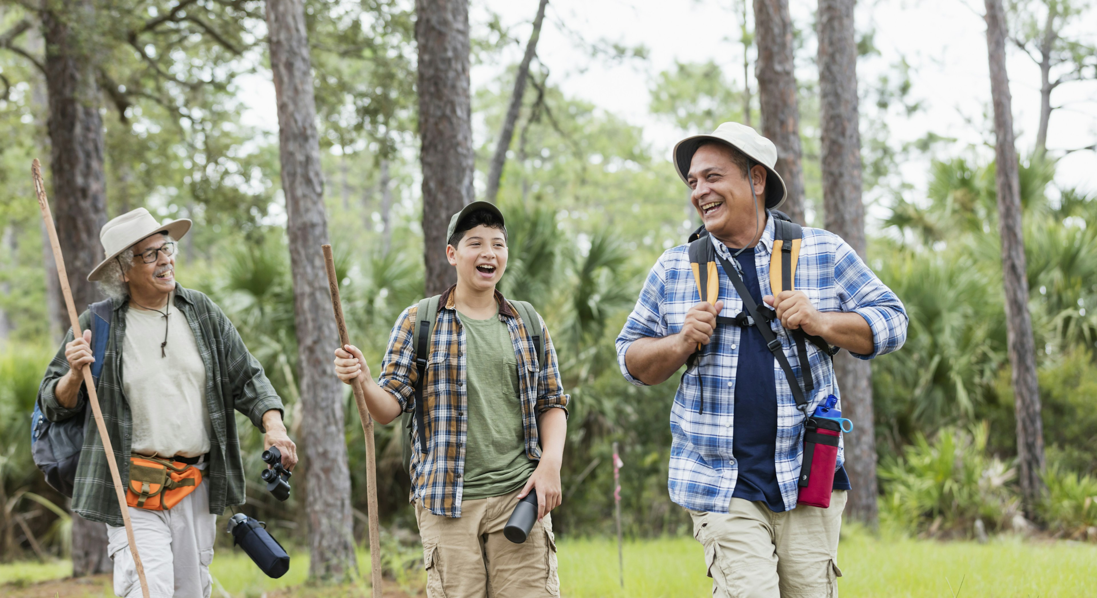 A multi-generation Hispanic family hiking together outdoors in the woods, wearing plaid shirts and hats and carrying walking sticks. The 12 year old boy walking in the middle, behind his father, a mature man in his 50s, and in front of his grandfather, a senior man in his 80s.
1129648546
hiking, healthy lifestyle, leisure activity, public park, natural parkland, woodland, nature, exploration, discovery, summer, springtime, outdoors, casual clothing, plaid shirt, hat, hiking pole, three people, senior men, mature men, boys, latin american and hispanic ethnicity, child, 12-13 years, 50-54 years, 80-89 years, vitality, active seniors, multi-generation family, father, son, grandfather, grandson, togetherness, bonding, friendship, smiling, happiness, cheerful, relaxation, candid, walking, talking, laughing, real people, waist up, florida - us state, adventure, retirement, sc0993