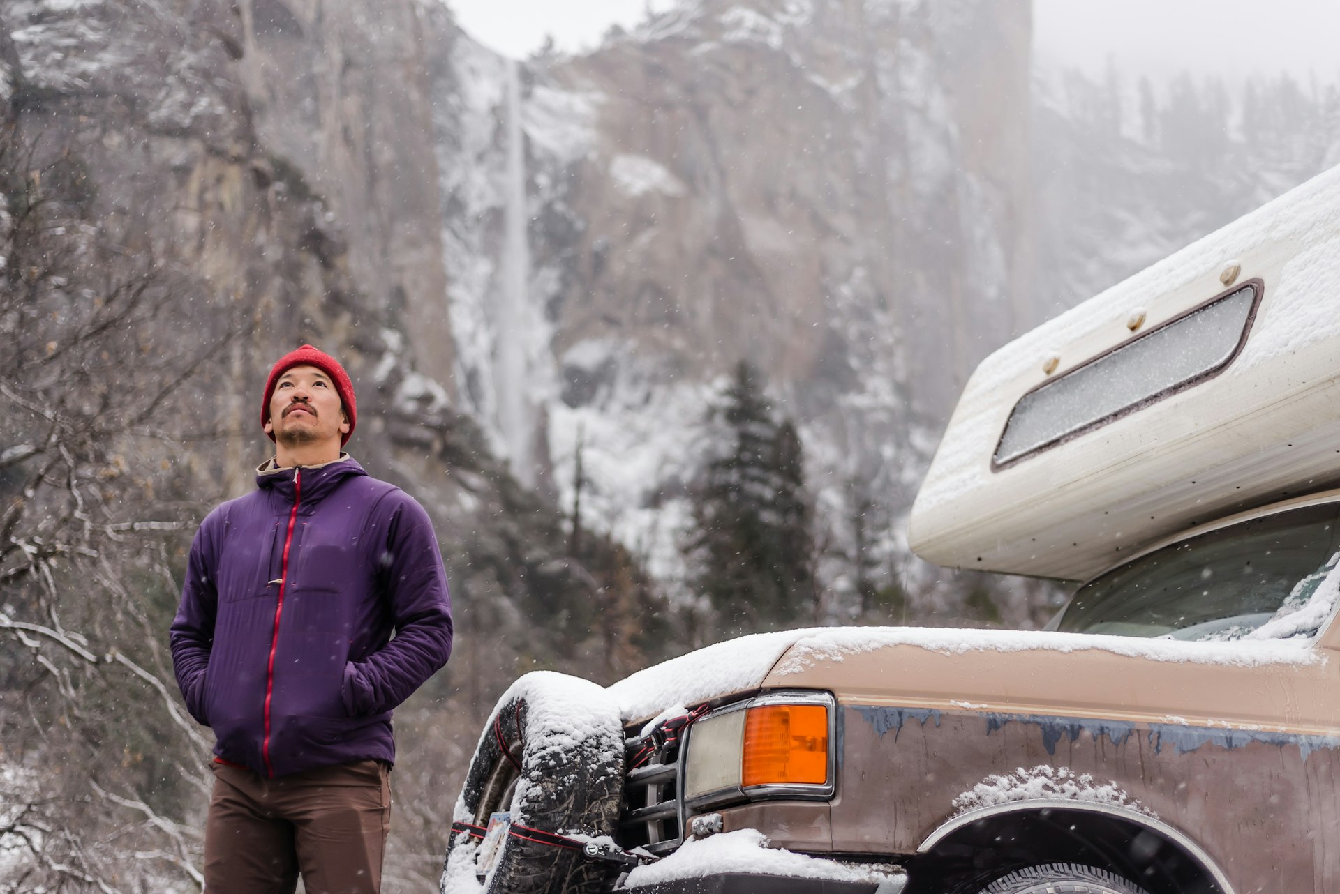 A man wearing snow gear stands in front of an icy waterfall next to an RV